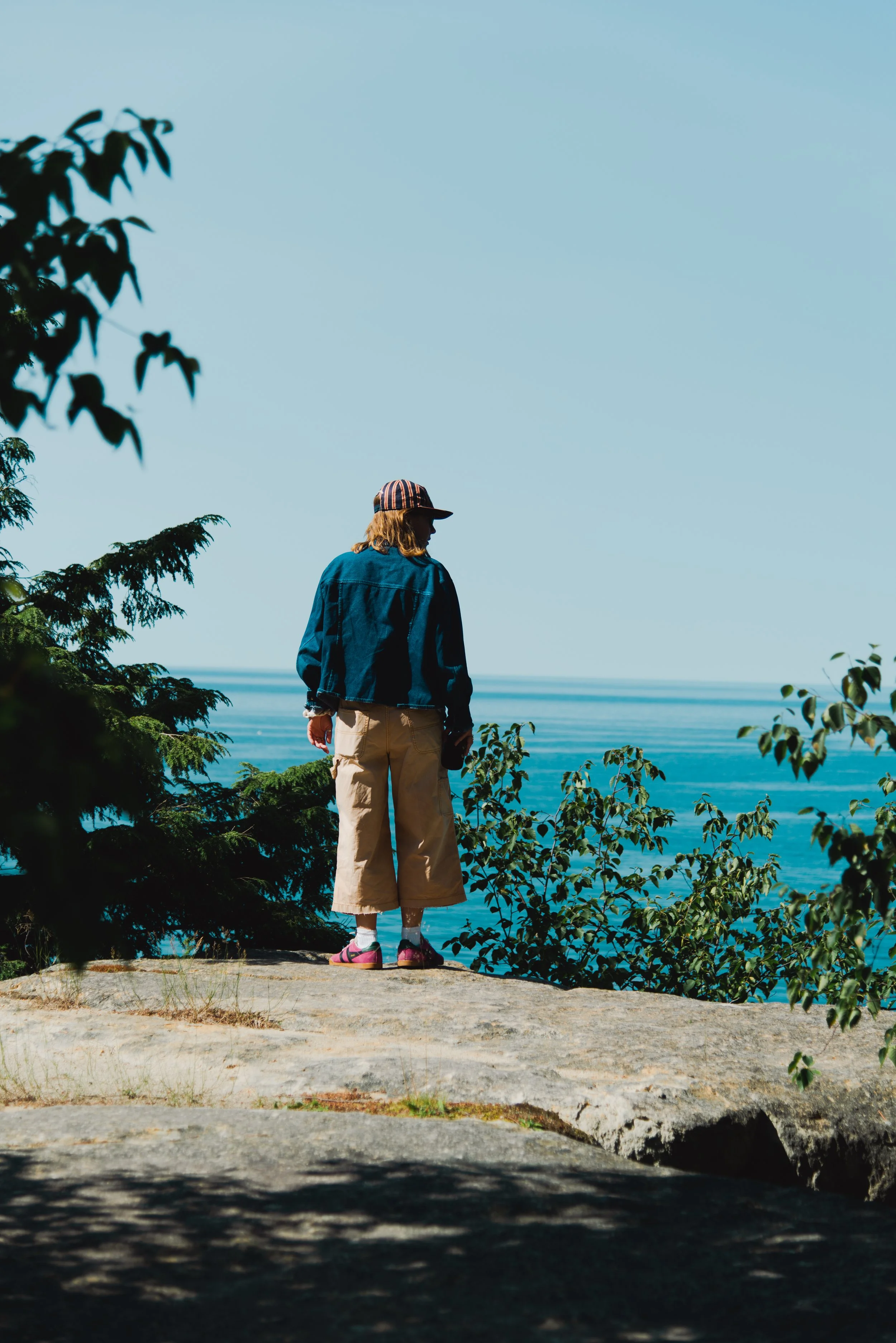 A person with long hair, wearing a striped baseball cap, a blue jacket, beige pants, and pink shoes, standing on a rocky area looking out over a body of water with trees around.
