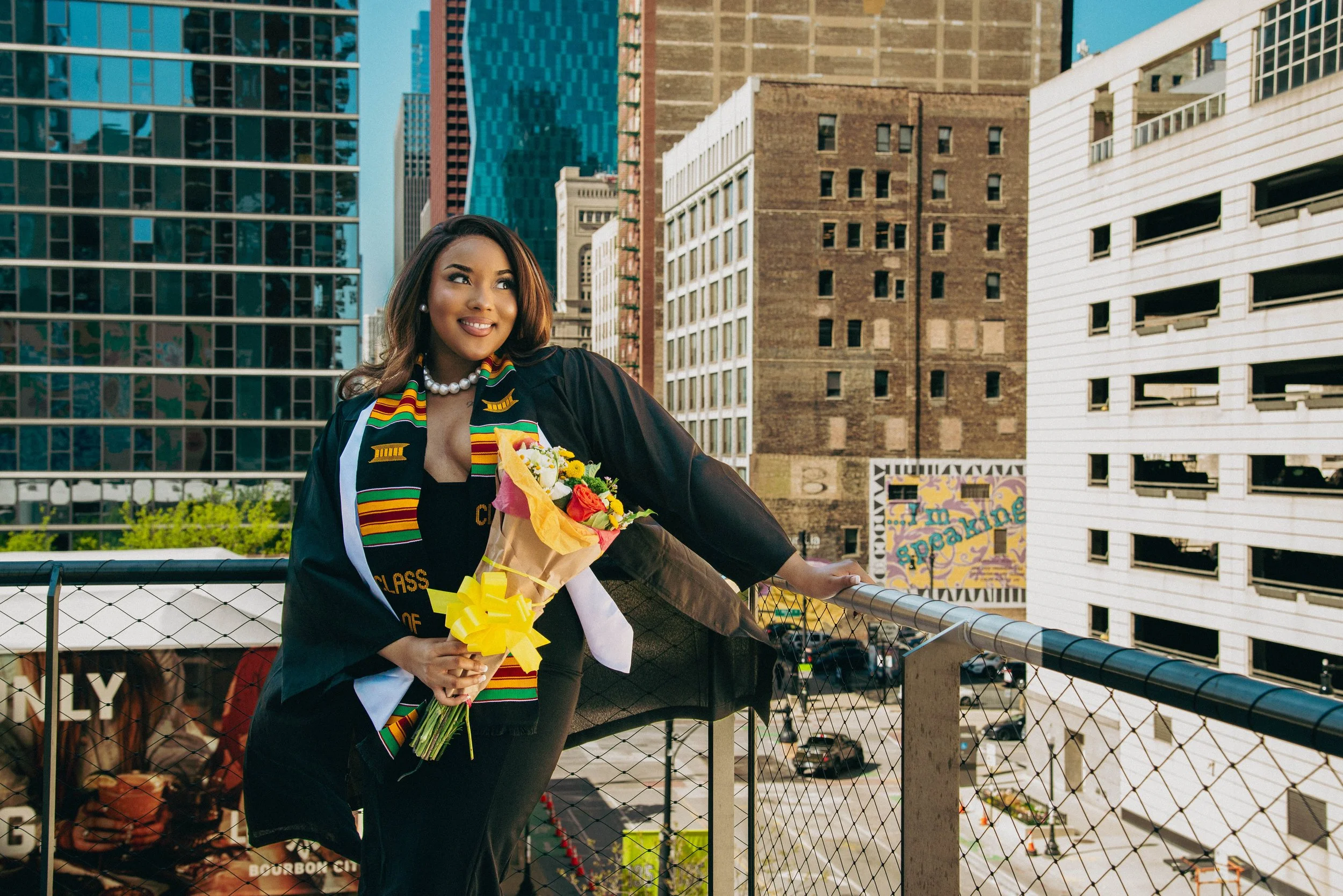 A woman in a graduation gown and sash holding a bouquet of flowers on a rooftop with city buildings in the background.