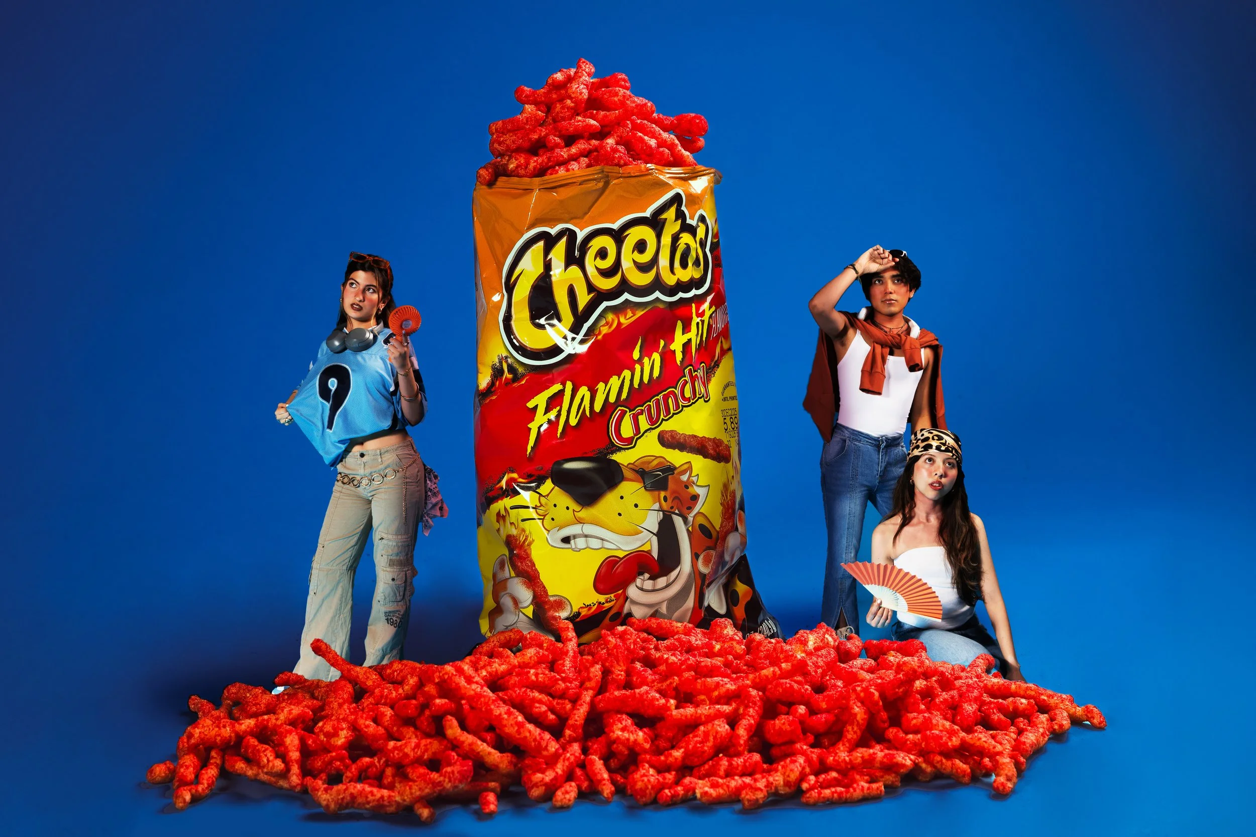 Four young women standing around a giant bag of Flamin' Hot Cheetos with some spilled on the floor. One woman is holding a fan, another is holding sunglasses, and one is wearing a headband. The bag has bright yellow, red, and black packaging and is s