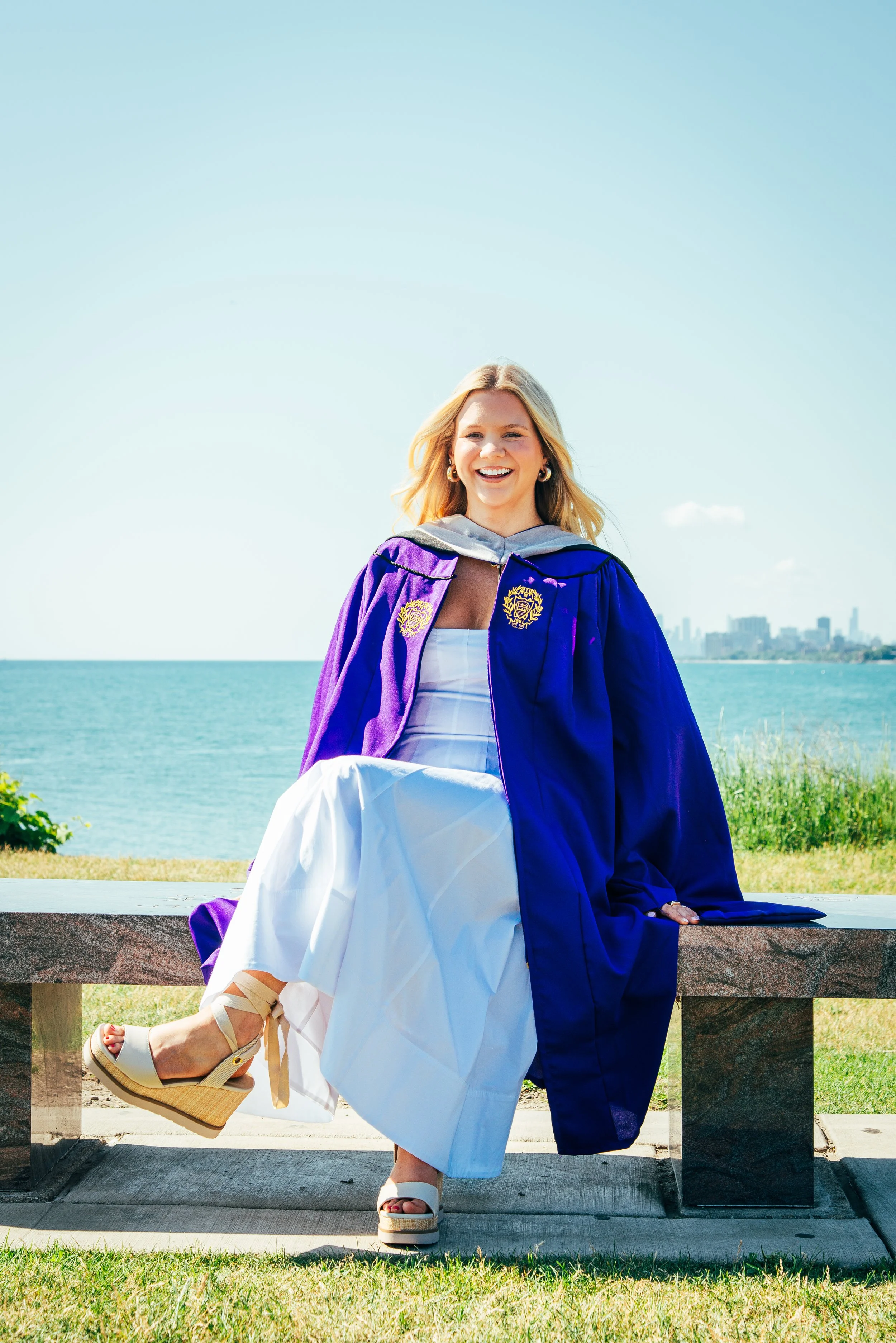 A young woman in a graduation cap and gown sitting on a bench outdoors near water, smiling, with city skyline in the background.
