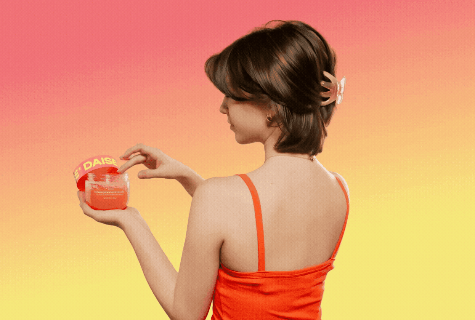 A woman with short brown hair wearing an orange dress, holding and looking at a small red container with a yellow lid that has the words 'E. DAISE' on it, against a gradient background of pink, orange, and yellow.