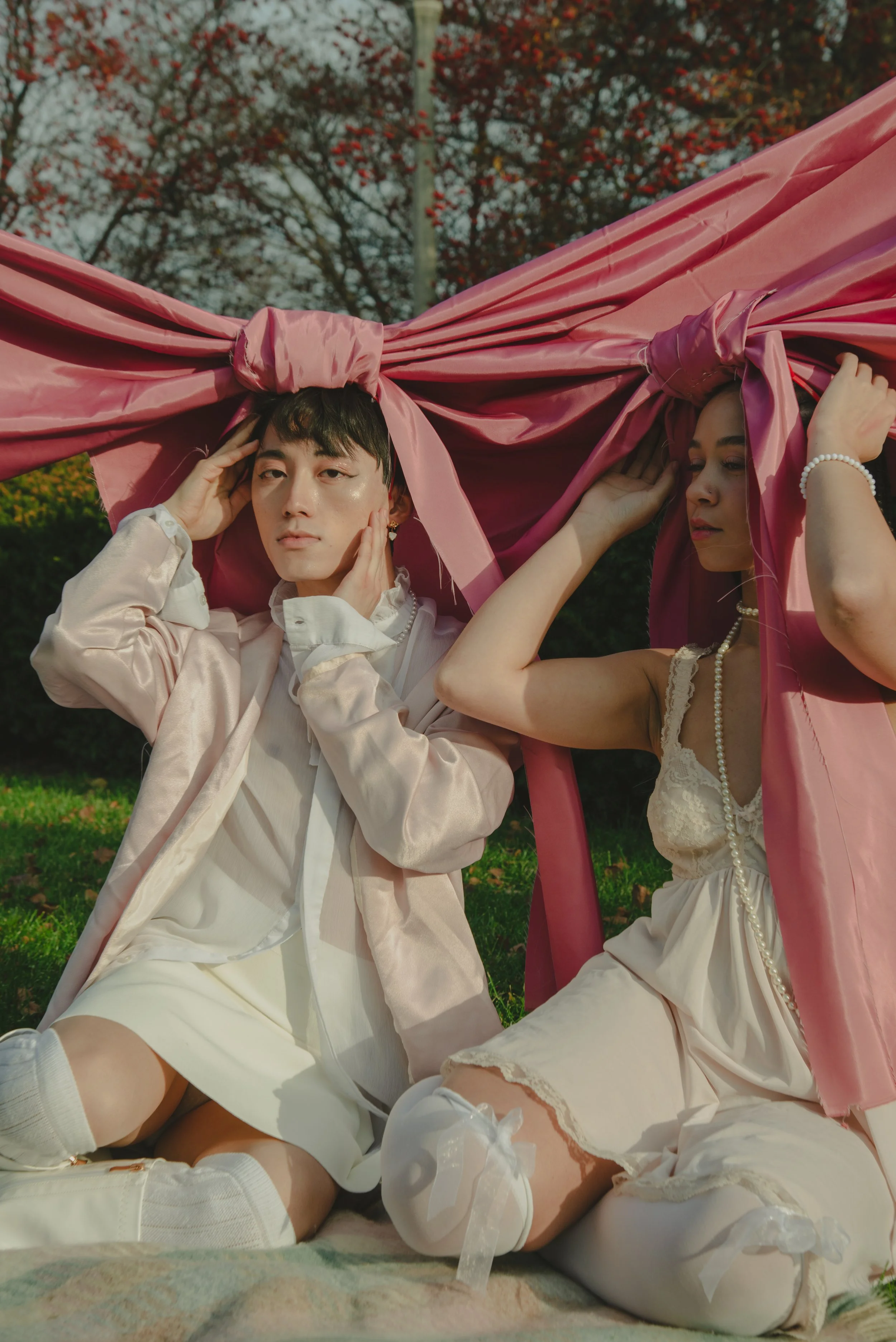 Two individuals dressed in vintage-style clothing, lying on the grass outdoors under a pink fabric canopy, with trees with red leaves in the background.