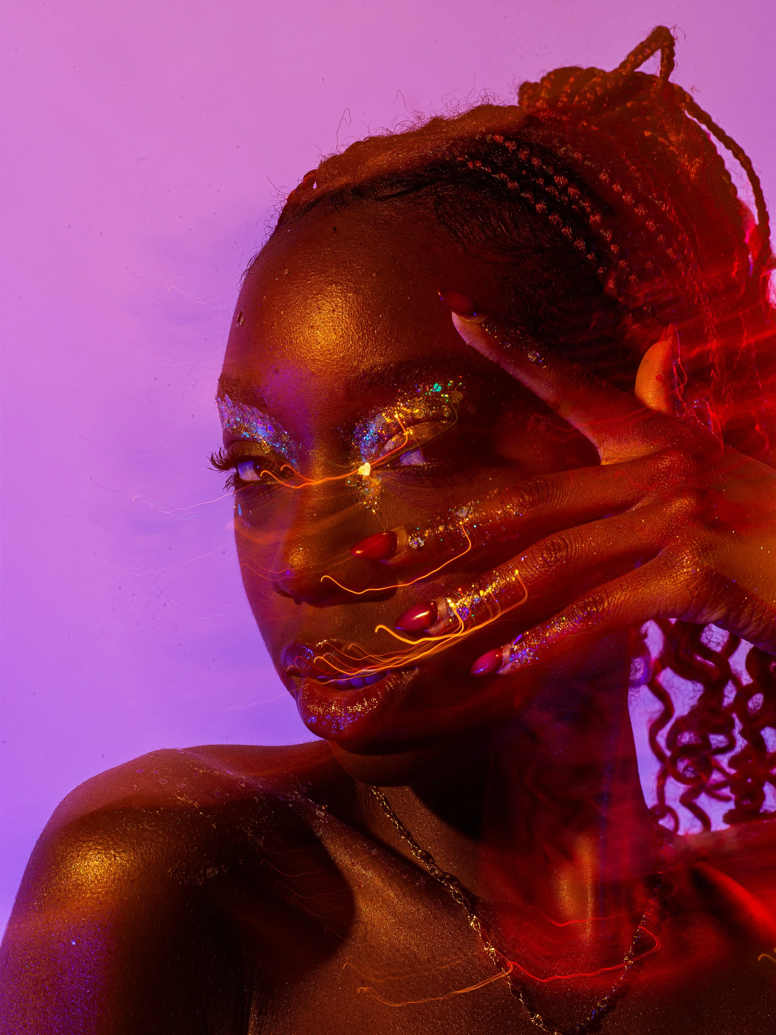 A person with dark skin and curly hair styled in braids is posing with their hand near their face. They have makeup with colorful glitter around their eyes and are illuminated with red, purple, and gold lighting, creating a vibrant, artistic effect.