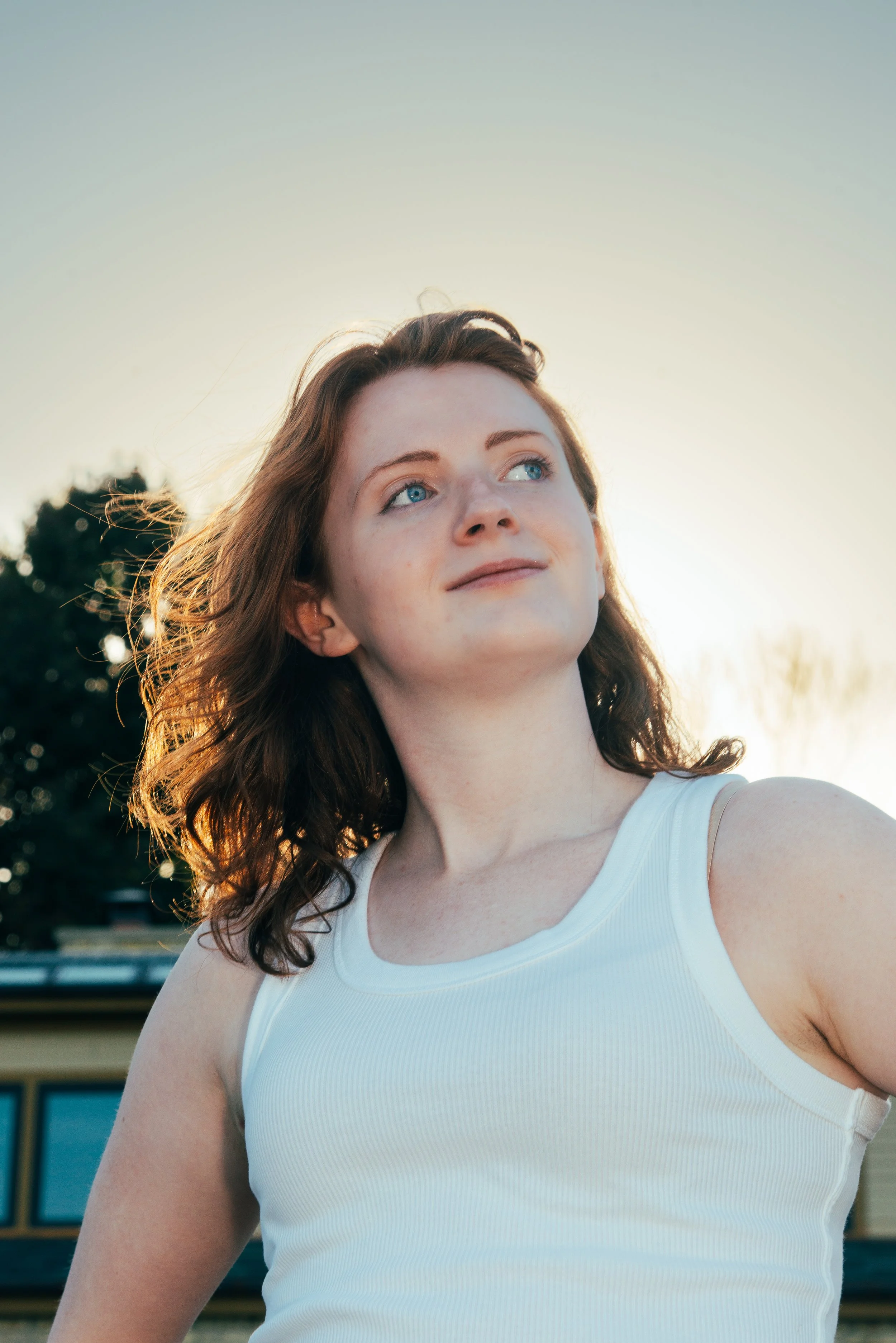 A young woman with wavy red hair and blue eyes smiling while looking to the side, wearing a white sleeveless top outdoors during daytime with a blurred background of trees and a building.
