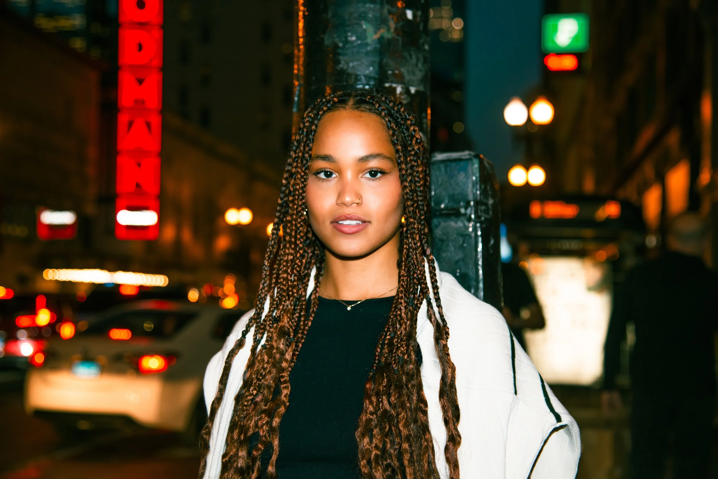 A young woman with braided hair and a nose ring standing on a city street at night, illuminated by streetlights and neon signs, with blurred cars in the background.