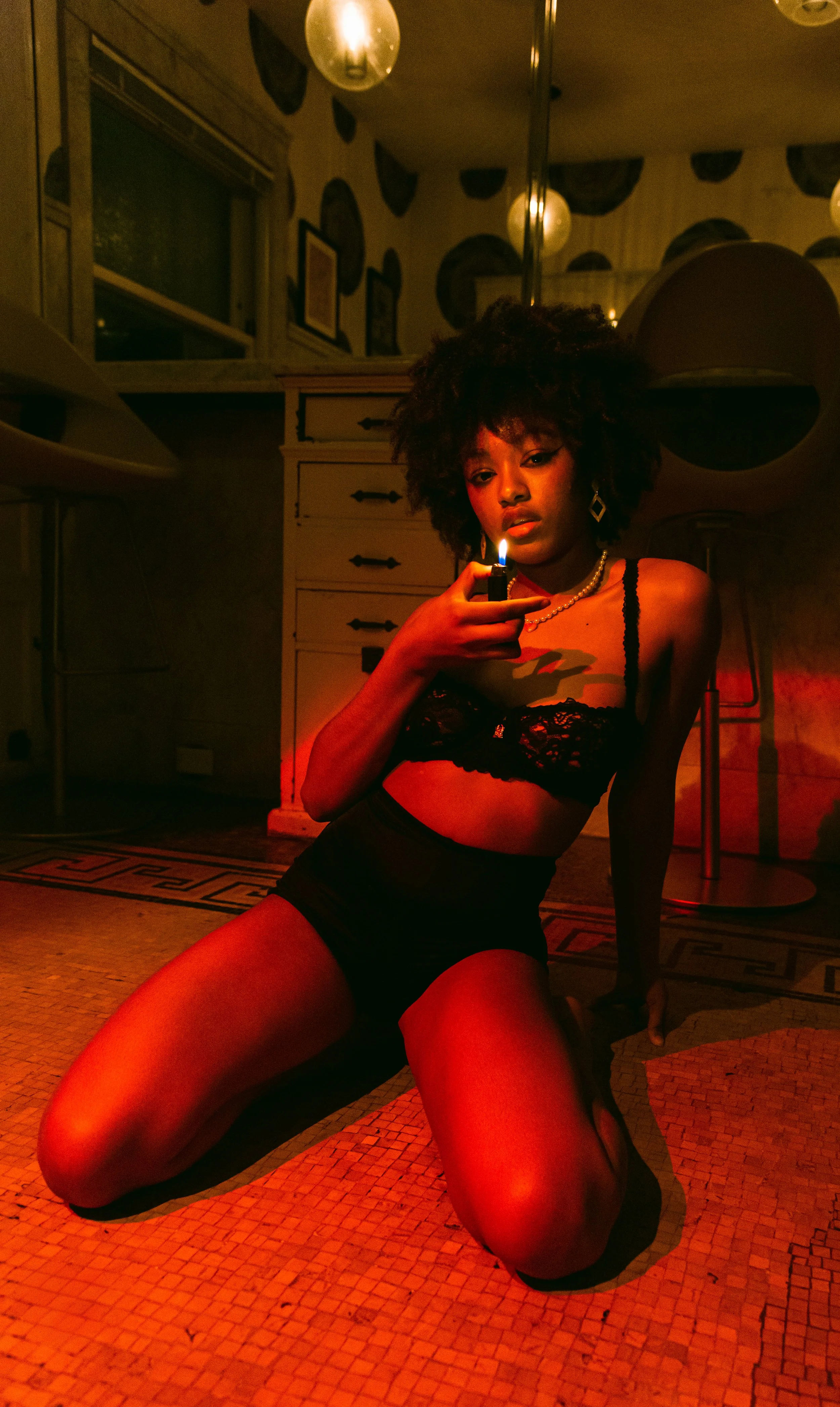 A woman with curly hair wearing black lingerie and high-waisted shorts, sitting on the floor in a dimly lit room, holding a lighter with a small flame.