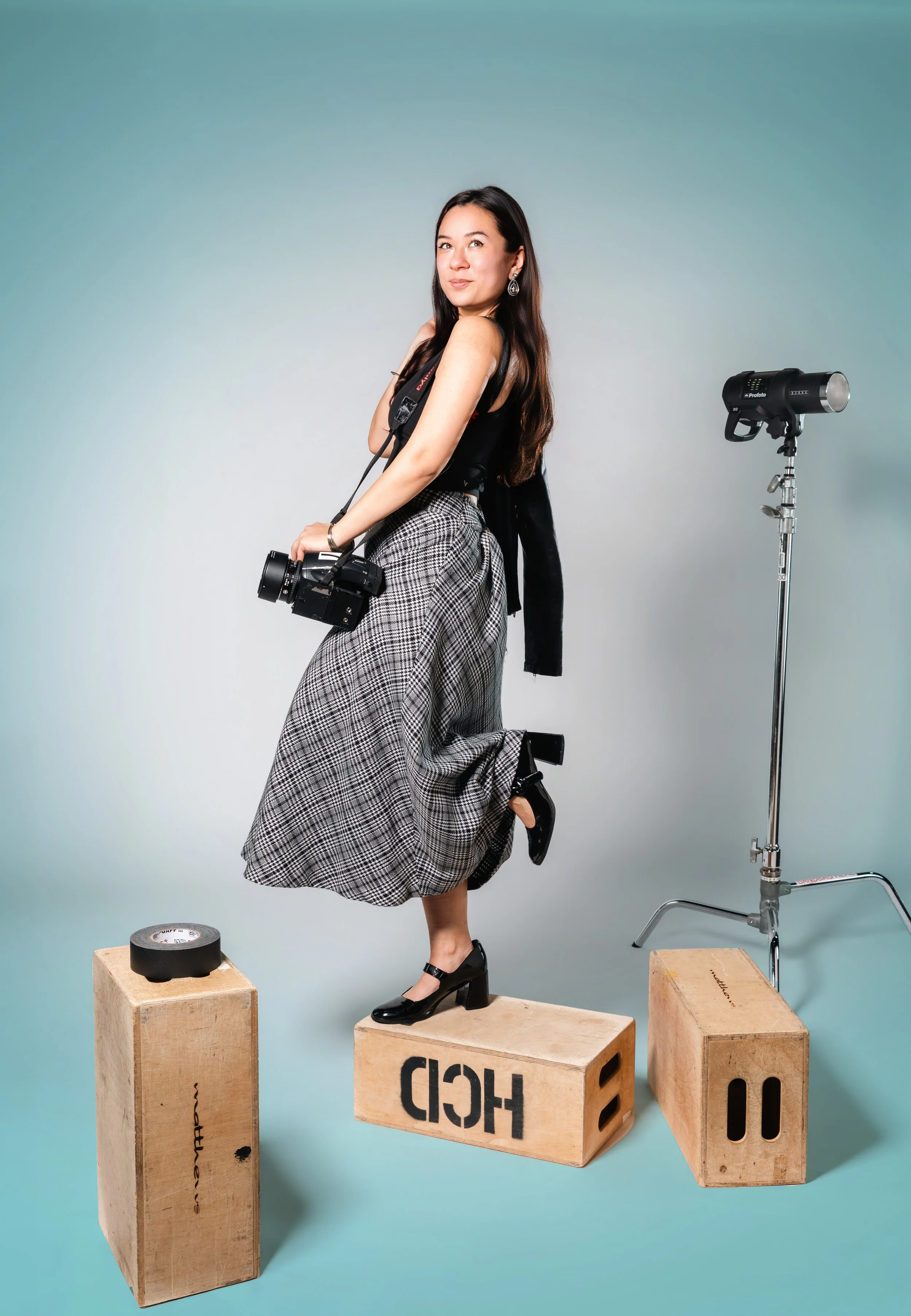 A woman with long dark hair and earrings stands on wooden blocks against a light blue background, holding a camera with a strap around her neck. She wears a black top, plaid trousers, and black heels. There is a studio light on a stand and a roll of black tape on a block nearby.