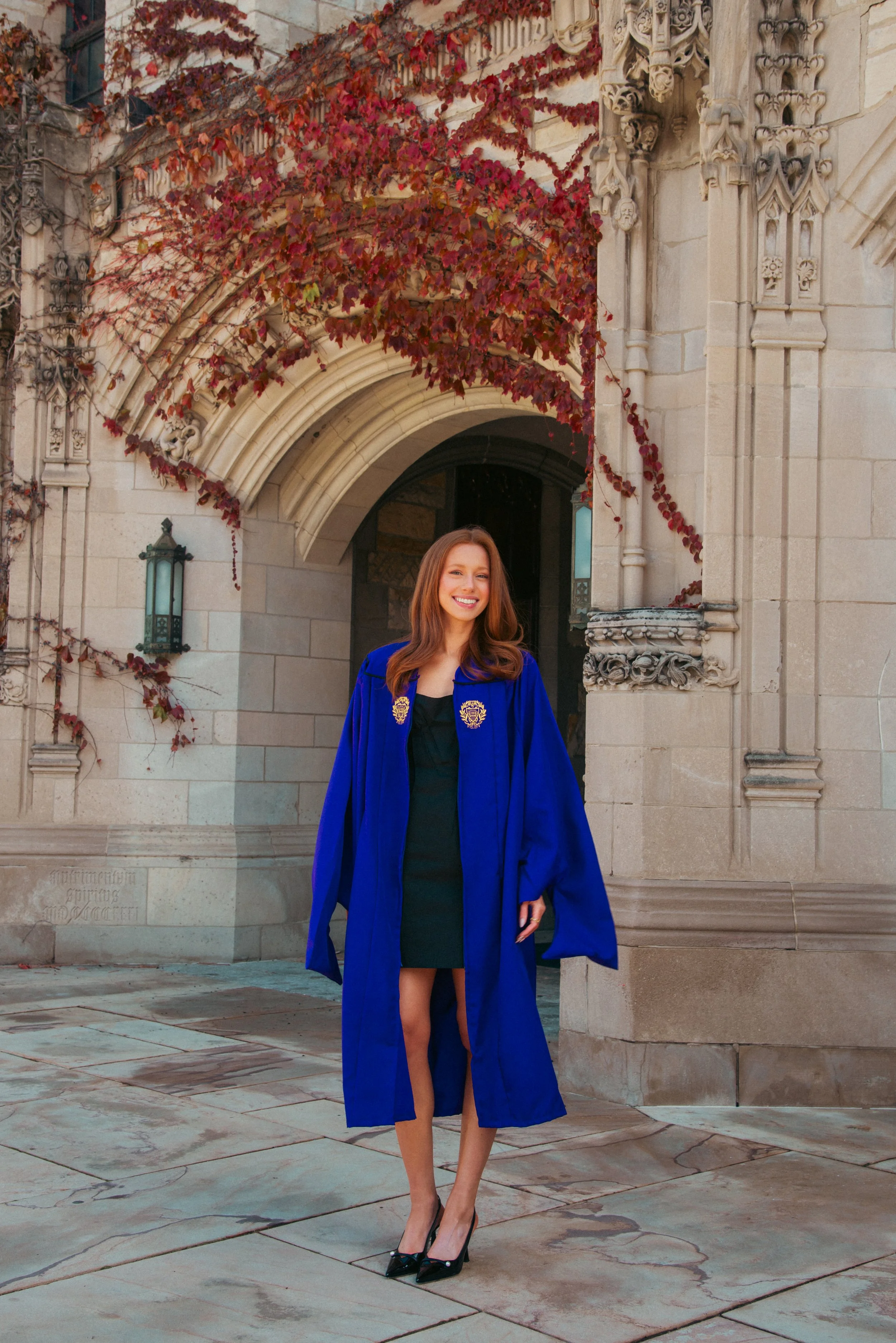 A young woman in a blue graduation gown and black heels standing in front of an ornate stone building with ivy-covered arches and Gothic architectural details. She is smiling and posing for the photo.