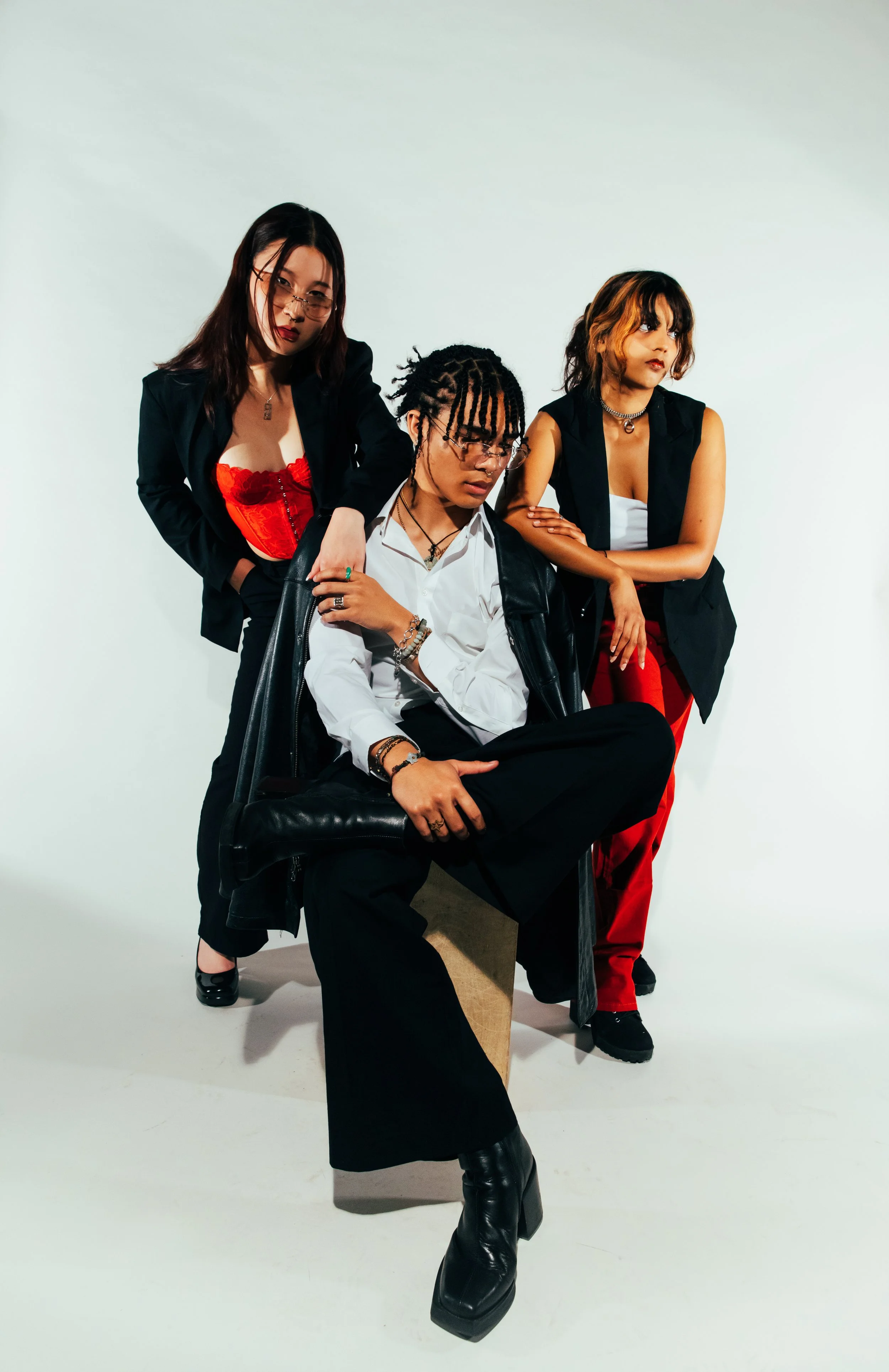Three young women with street style clothing posing against a plain white background. The woman in the center has braids and is sitting on a chair with one leg crossed over the other. The woman on the left has shoulder-length dark hair, glasses, and 