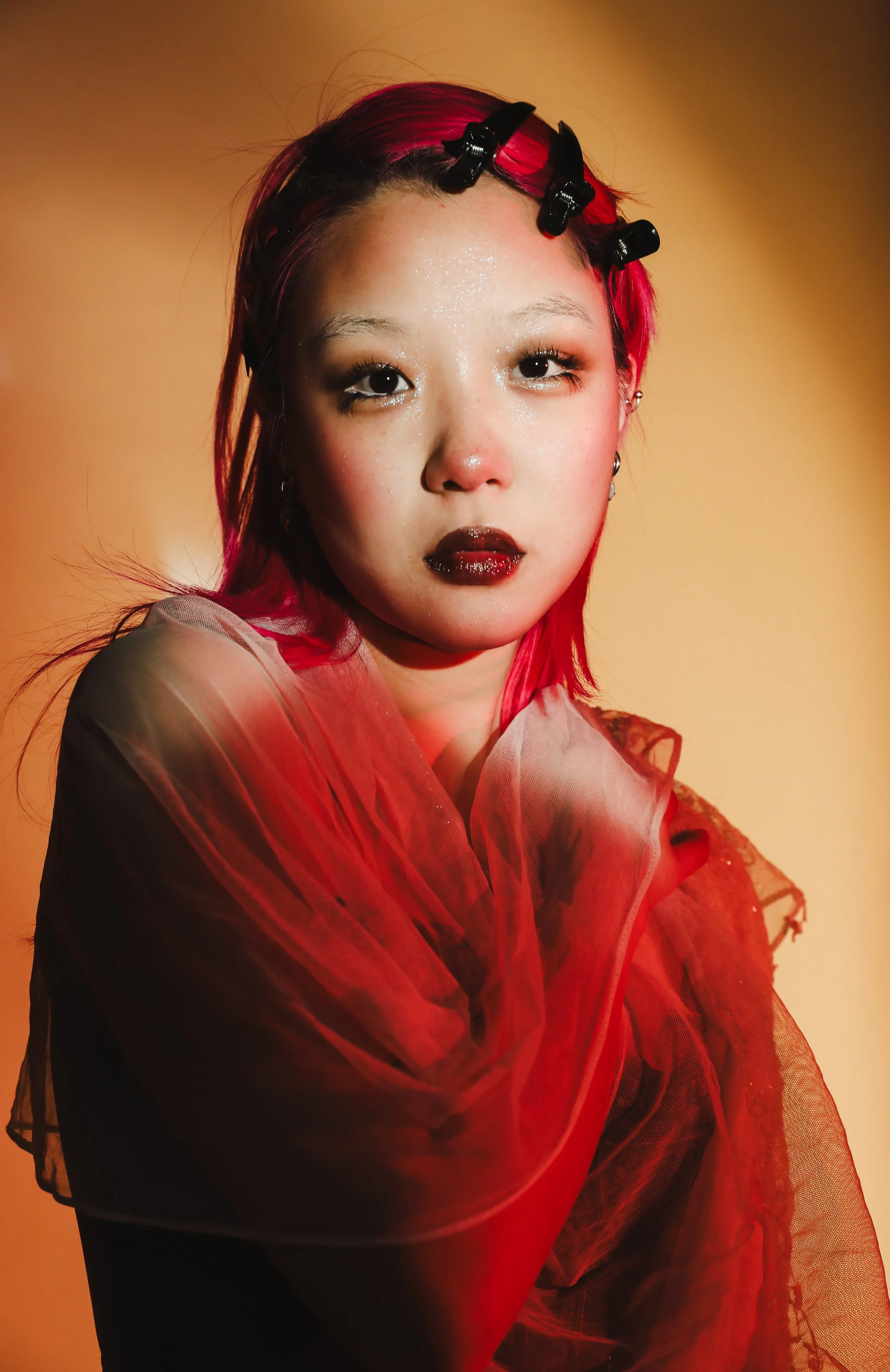 A woman with bright pink hair wearing black hair clips, dark lipstick, and shimmering eye makeup, draped in sheer red fabric, standing against a warm orange background.