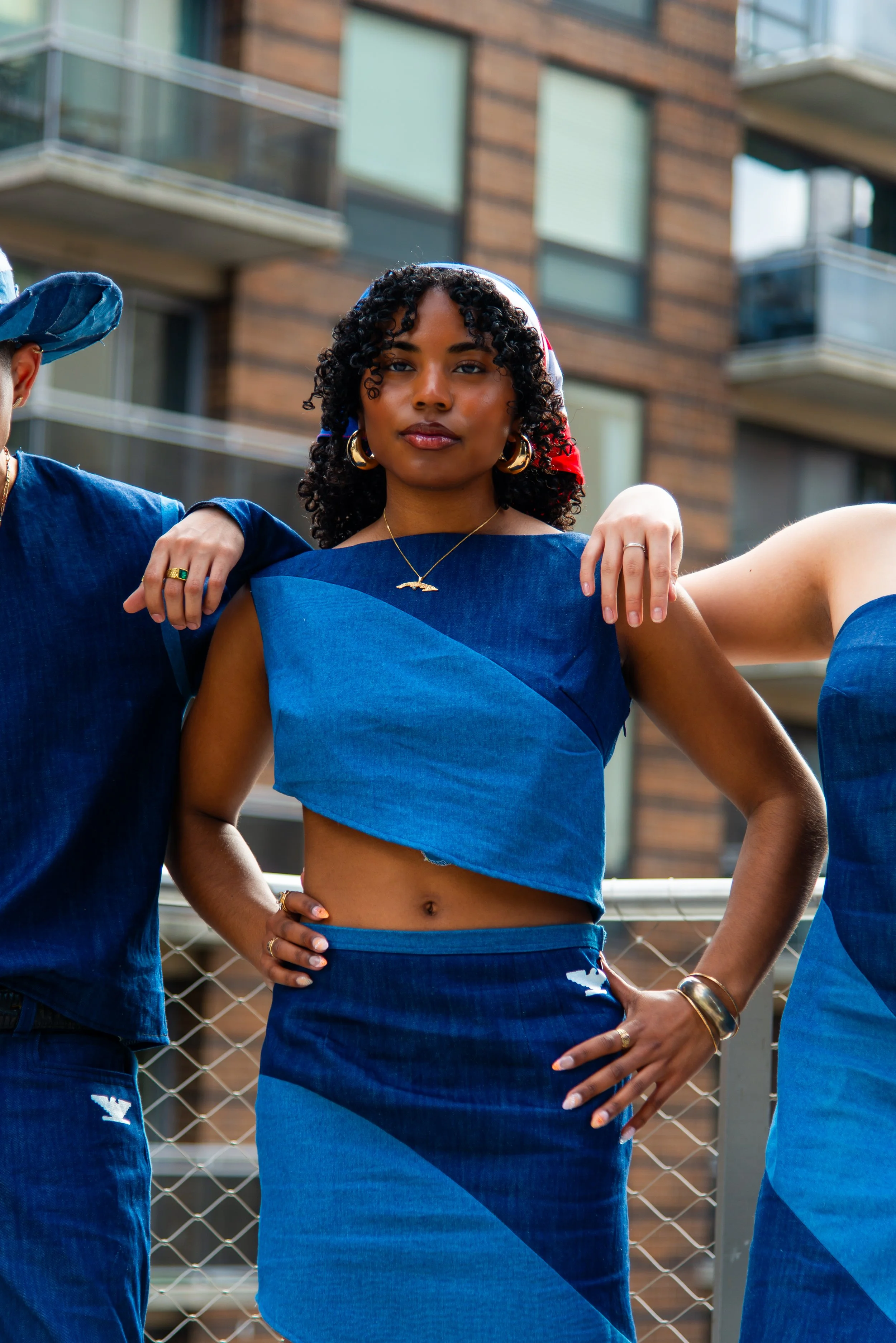 A woman with curly hair wearing a blue coordinated outfit, gold jewelry, and a white, red, and blue bandana, standing outdoors in an urban setting with apartment buildings in the background.