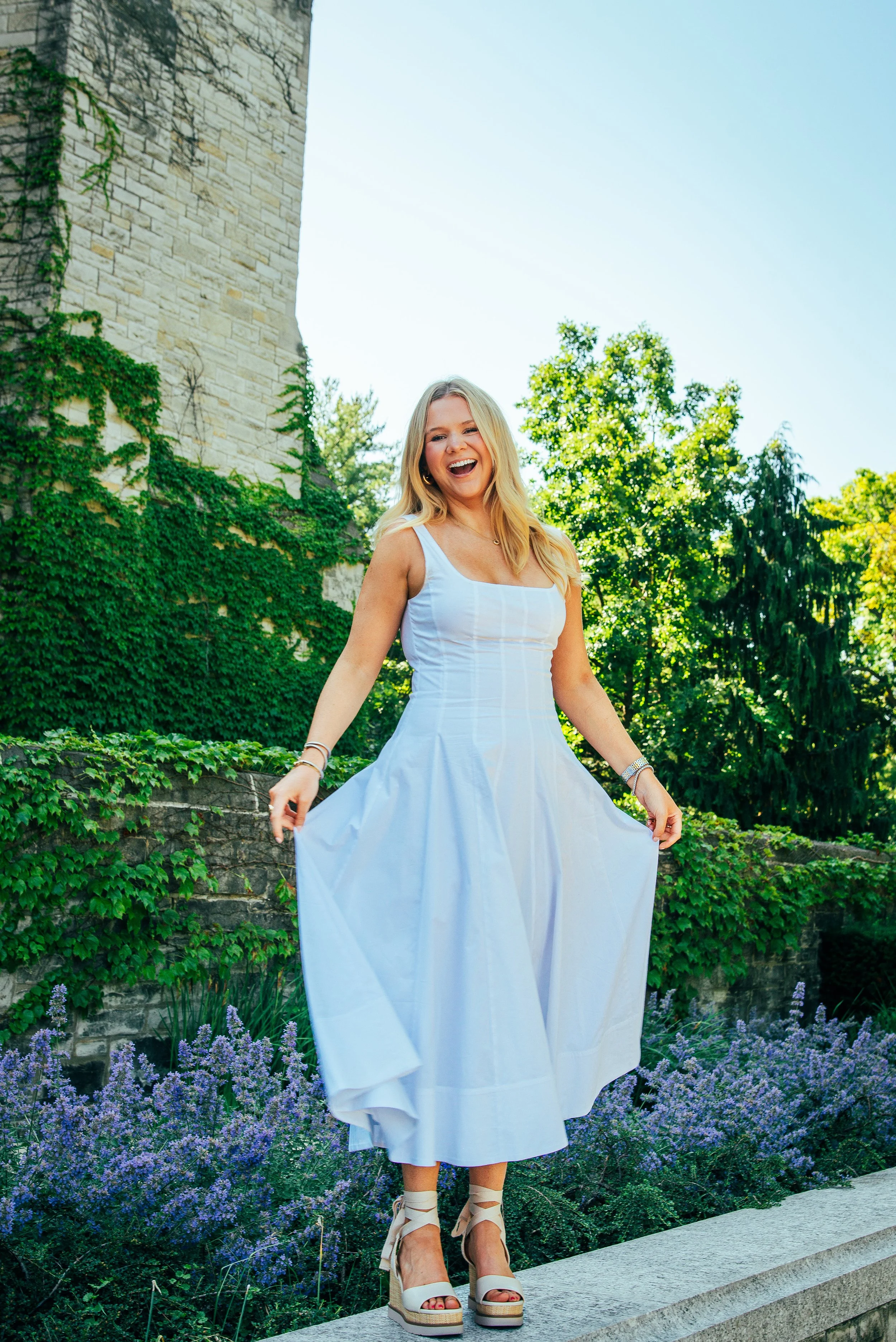 A woman wearing a white dress and high-heeled sandals standing outdoors near purple flowers and greenery, smiling and laughing.