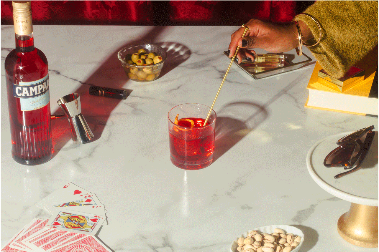 A marble table holds various items including a bottle of Campari, a jigger, playing cards, a bowl of olives, a cocktail in a glass with a stir stick, a pair of sunglasses on a plate, a container of pistachios, a stack of books, a small mirror, and a 