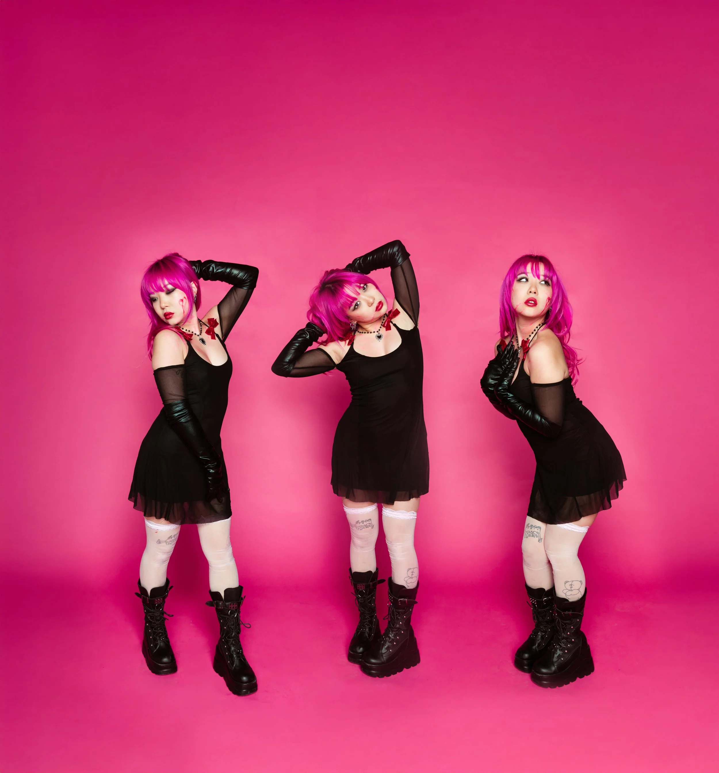Three women with bright pink hair standing against a pink background. They are wearing black dresses, black gloves, and black boots, with white stockings featuring tattoos and small designs.