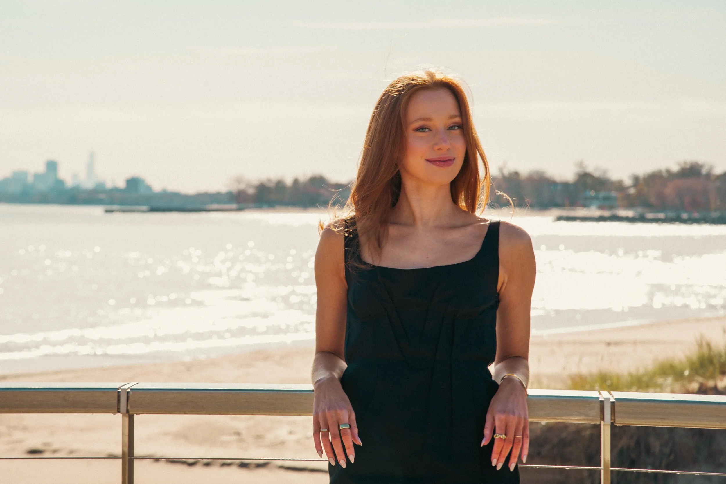 A woman with red hair wearing a black dress, standing in front of a body of water with a city skyline in the distance on a sunny day.