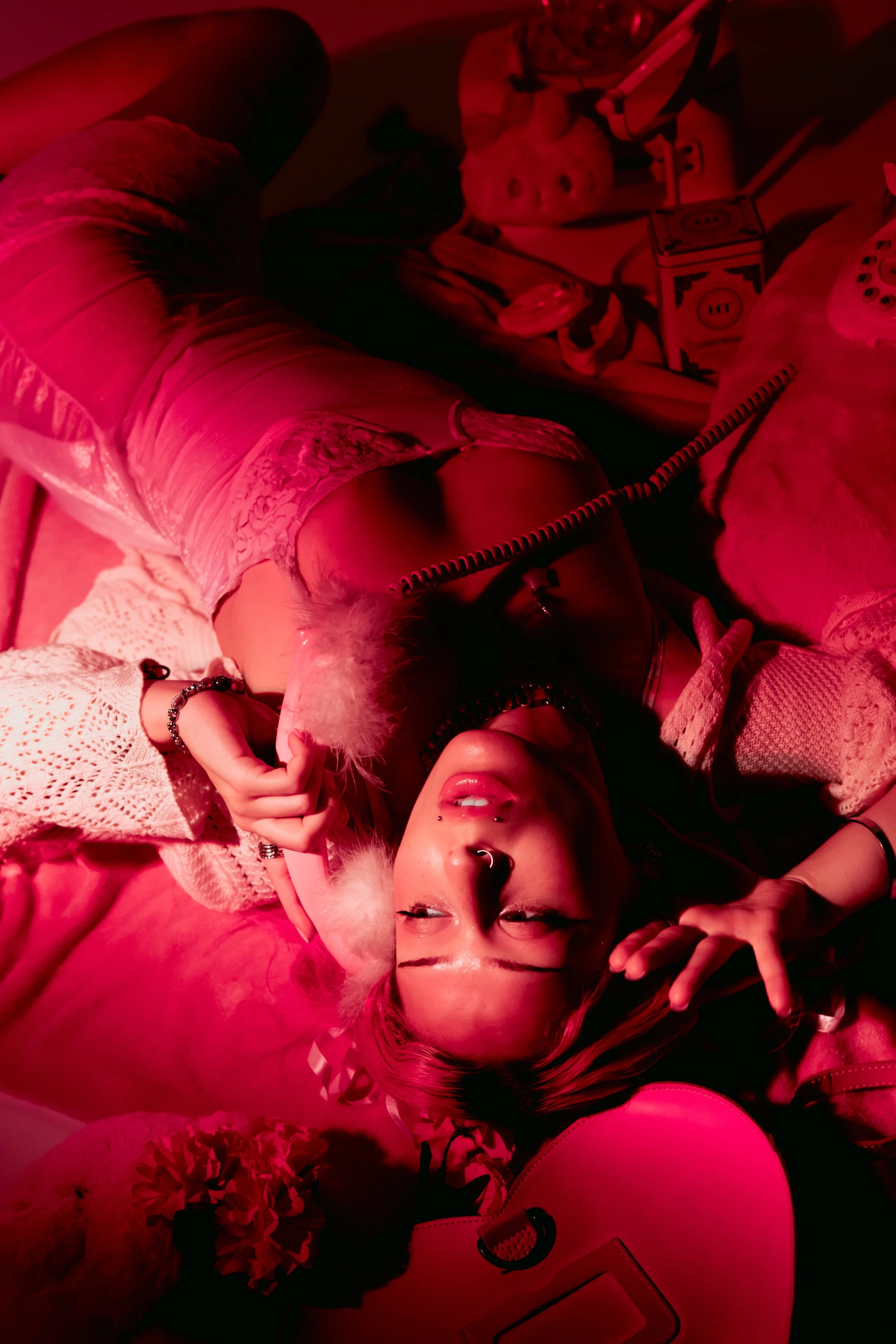 Person with dyed pink hair lying on a bed, making a peace sign with their hand. They are wearing jewelry, including rings and bracelets, and have piercings on their face. The room is illuminated with red lighting and has various objects, including a 
