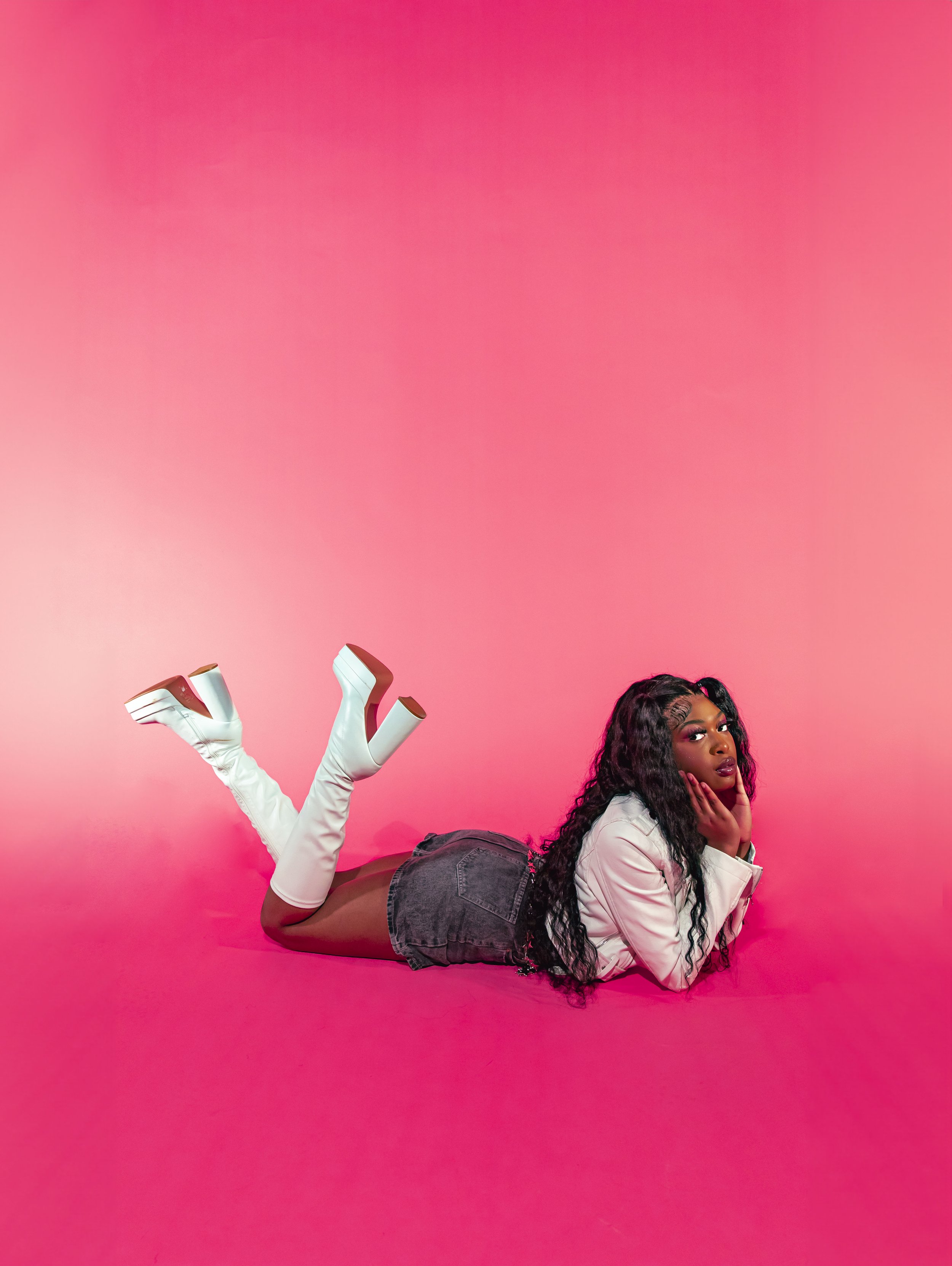 A woman with long curly black hair wearing a white jacket, denim shorts, and white knee-high boots, lying on her side against a pink background. She is resting her head on one hand and looking into the camera.