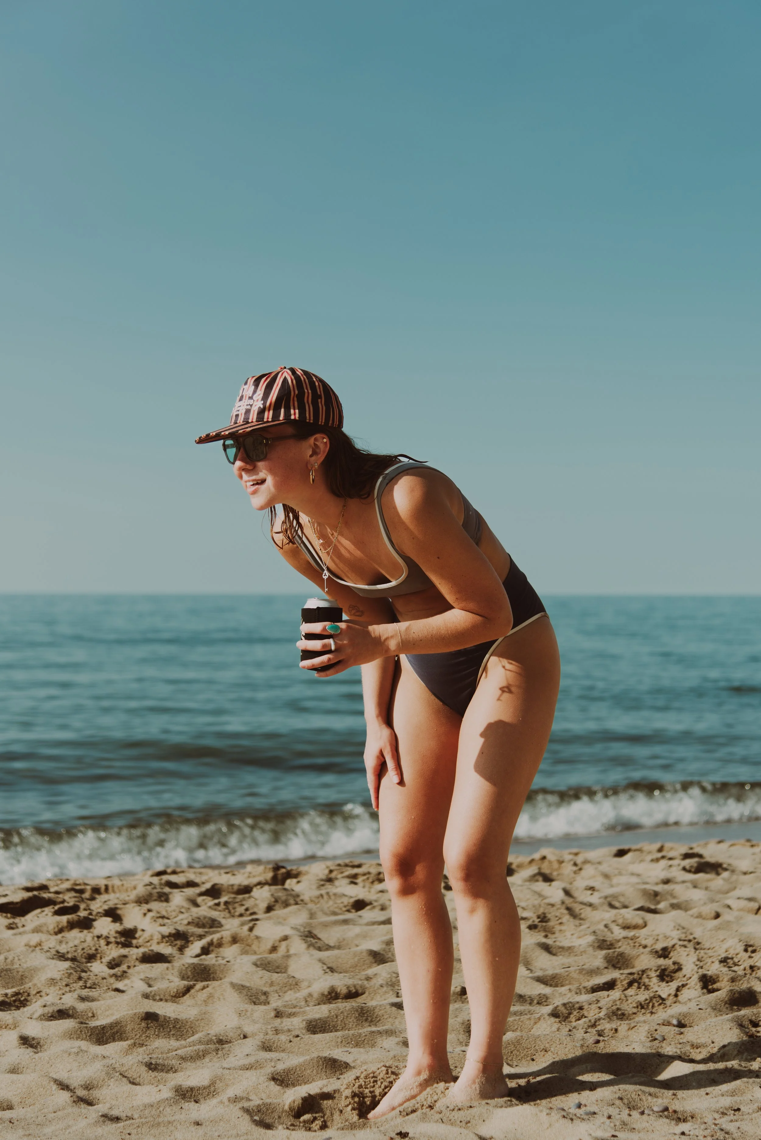 Woman in bikini and sunglasses on the beach holding a drink, bending over slightly, with the ocean and sky in the background.