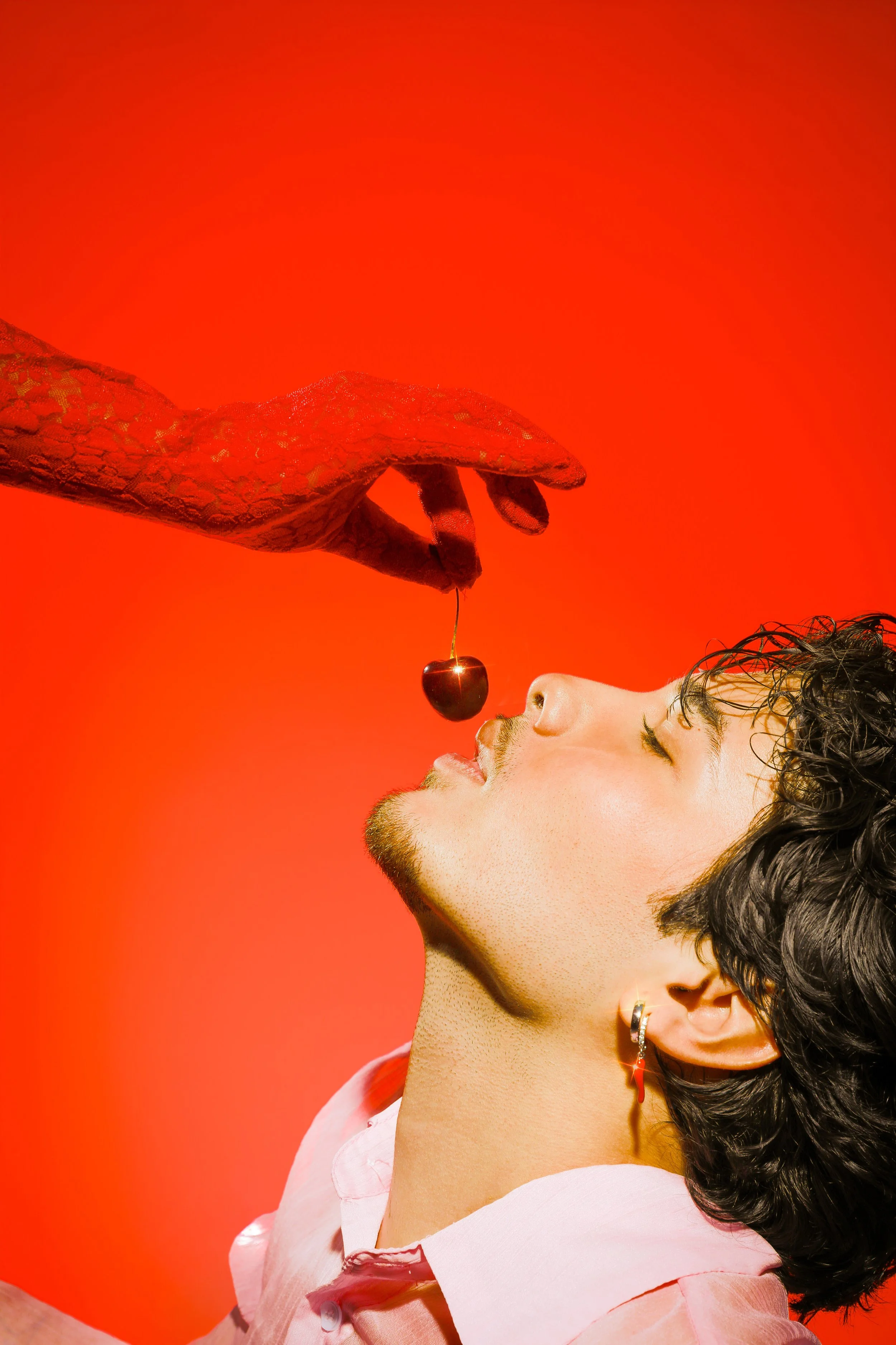 A young woman with dark, wavy hair and earrings is shown in profile against an orange background. She is wearing a light pink shirt and has her eyes closed, with a cherry being held by a hand near her mouth, about to be eaten.