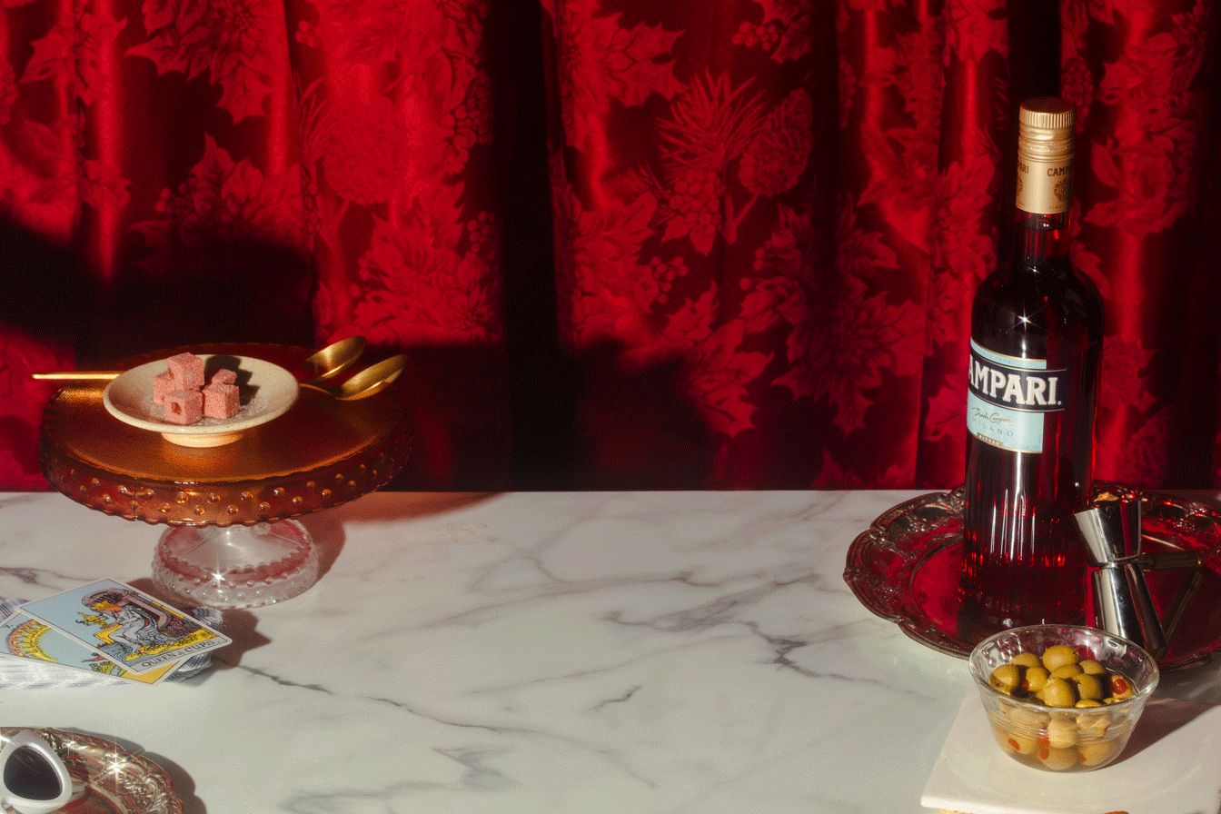 A marble tabletop with a bottle of Campari bitters, a bowl of green olives, a tarot card, a decorative cake stand with pink fudge, and gold spoons. Red velvet curtain with floral pattern in the background.