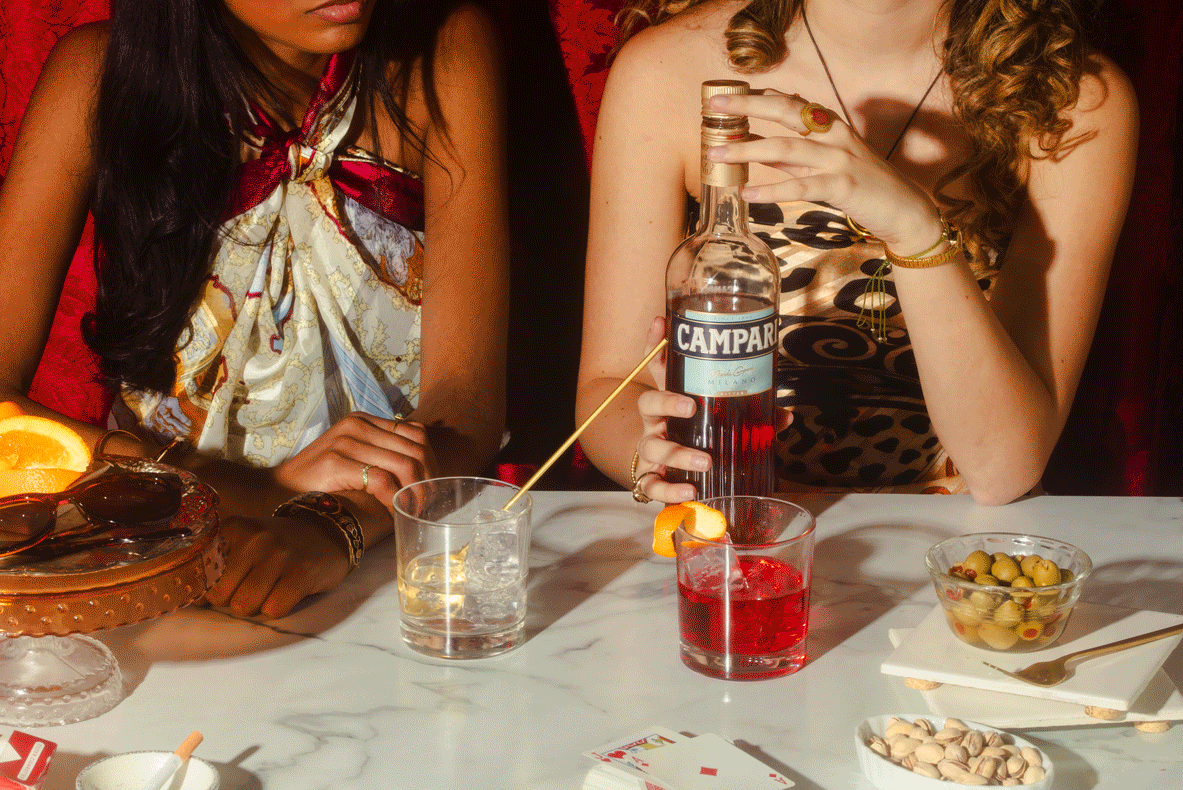 Two women sitting at a table with drinks and snacks, one holding a bottle of Campari and the other with a skewer and slice of orange on her drink