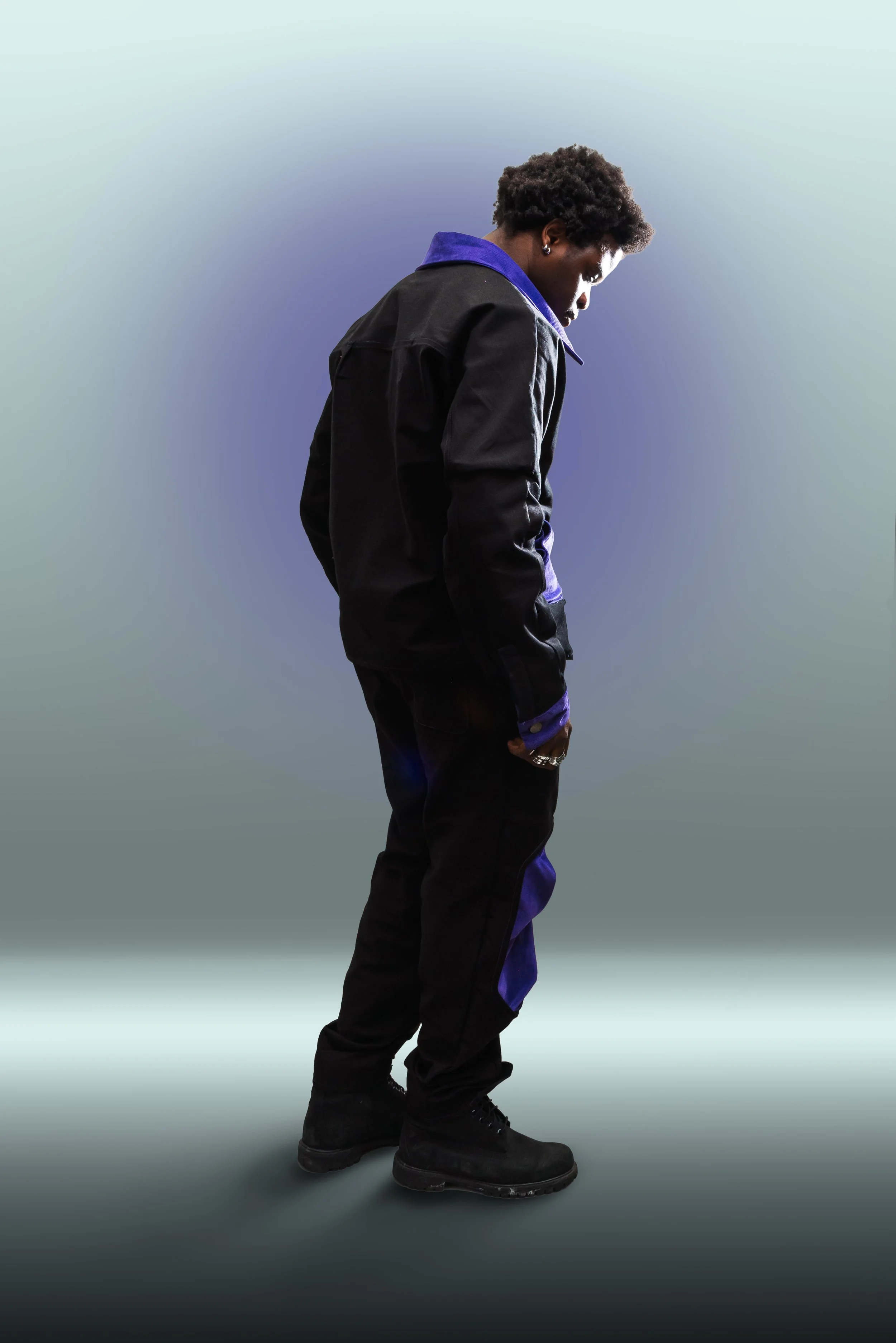 A Black man with curly hair wearing a black jacket with purple accents, black pants, and black boots, standing in front of a light gradient background with his head bowed and hands in his pockets.