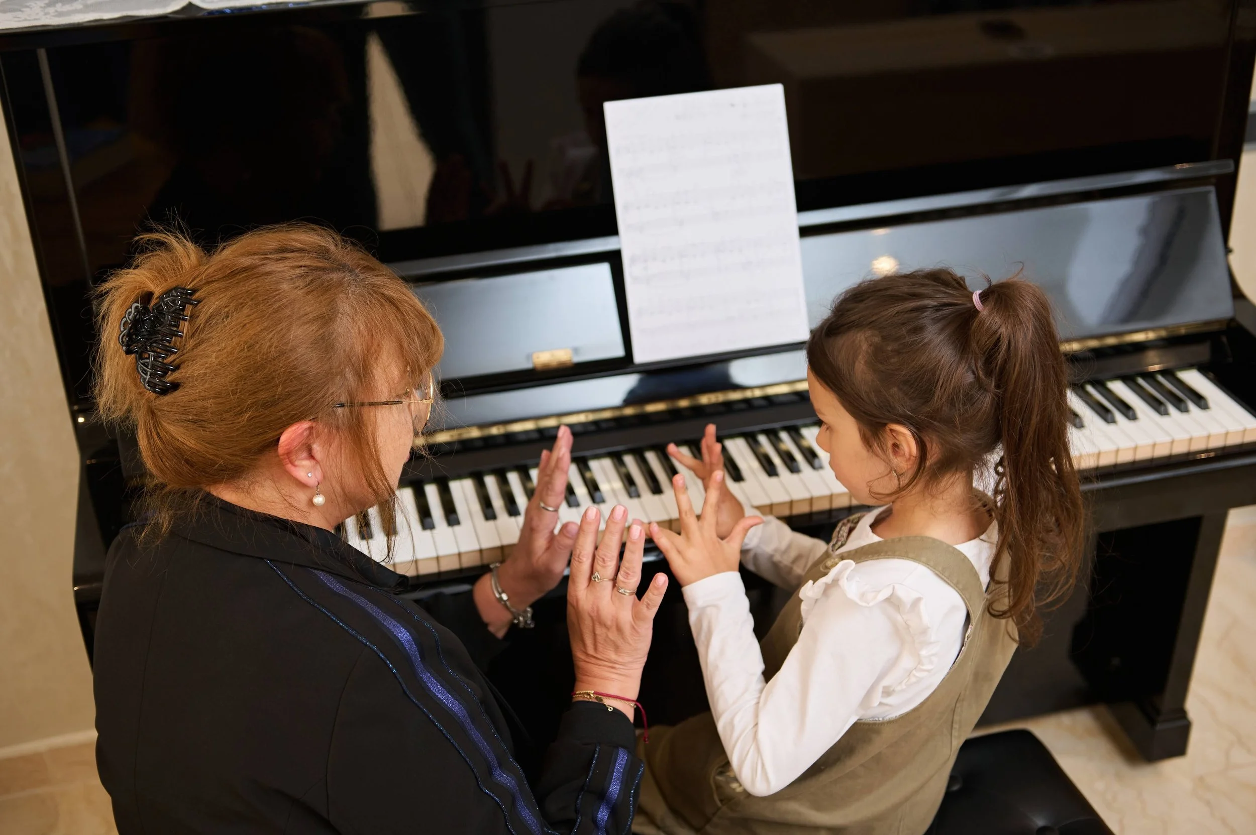 An adult woman and a young girl sitting at a piano, with the woman teaching the girl how to play, in a close-up view from above.