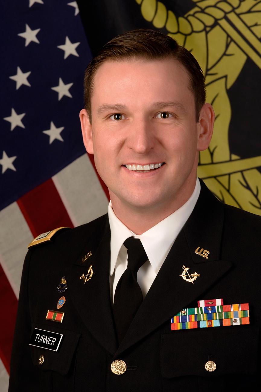 A young male military officer in uniform standing in front of American and military flags.
