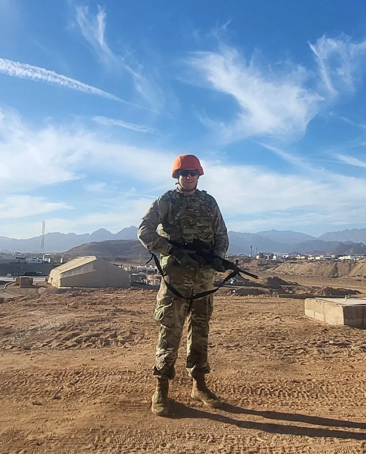 A soldier in camouflage uniform, red helmet, and sunglasses standing in a desert landscape with mountains and military structures in the background.