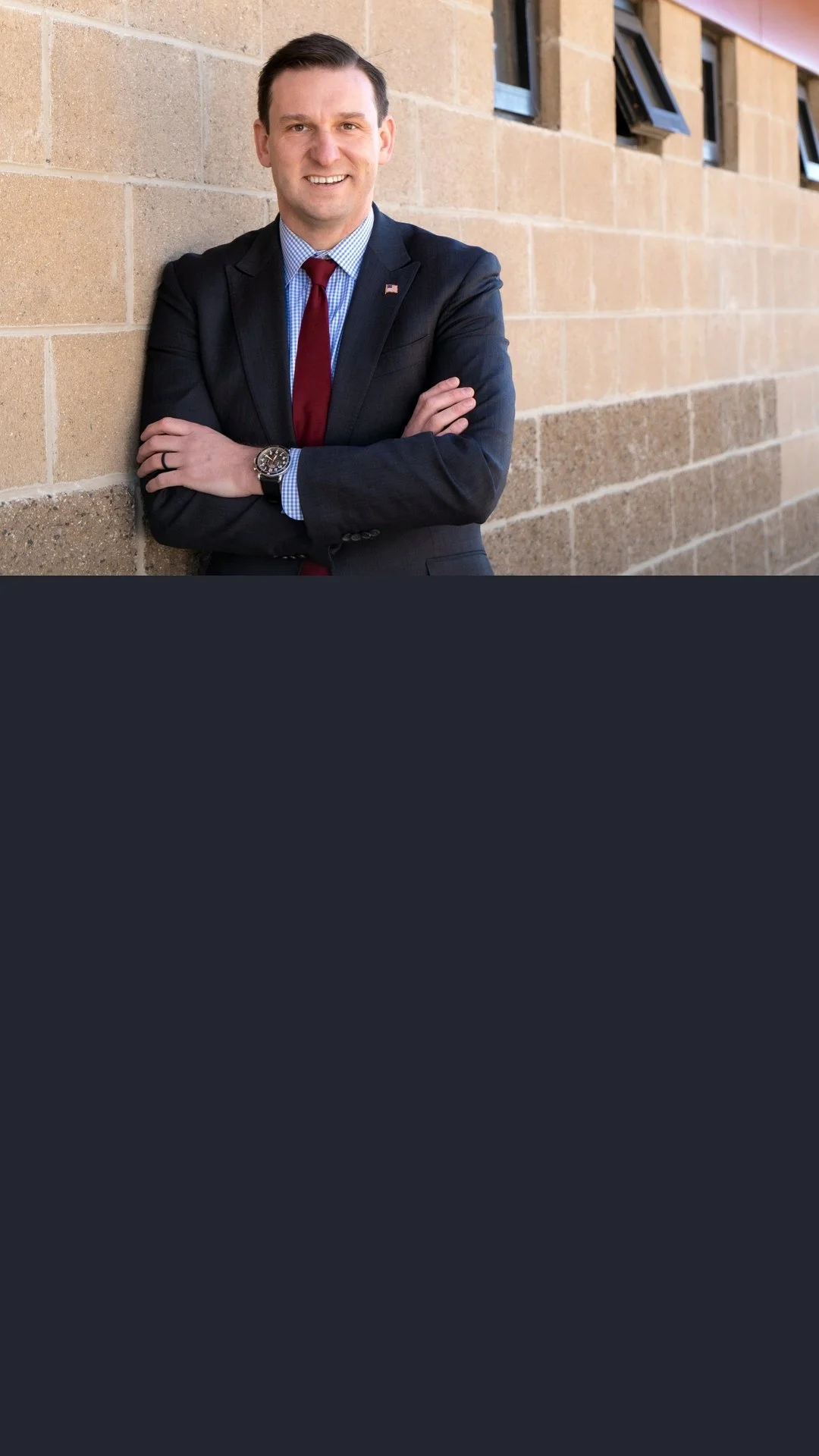 A man in a dark suit with a red tie, standing with arms crossed and smiling, leaning against a brick wall with small windows.