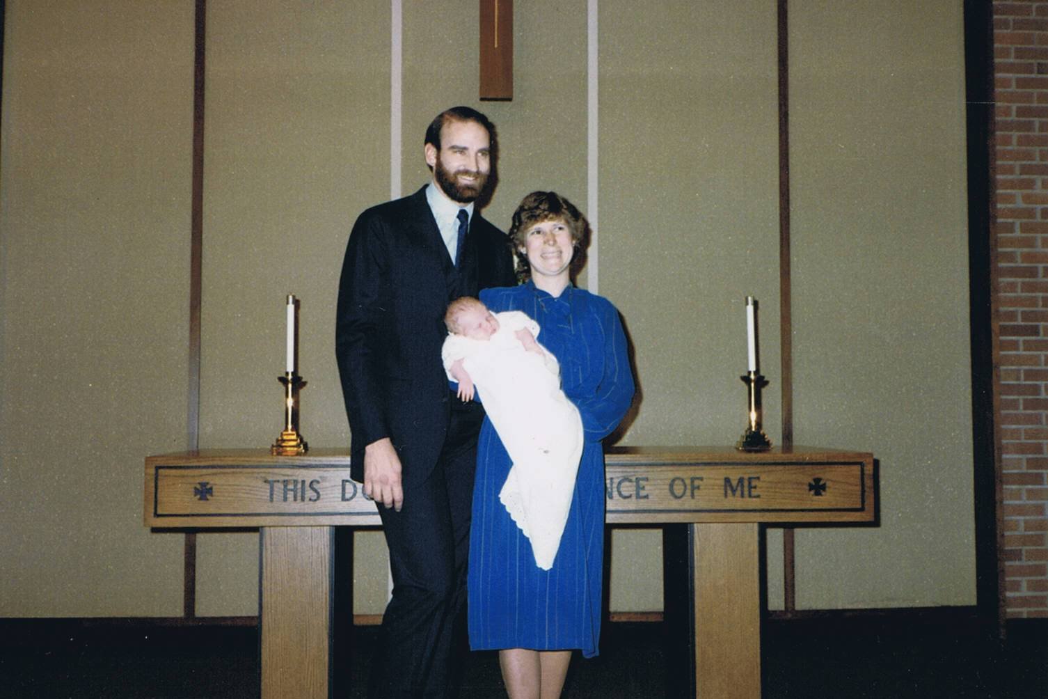 A family standing in front of an altar inside a church, with a man in a dark suit, a woman in a blue dress holding a newborn baby, and a backdrop with candles and a wooden cross.