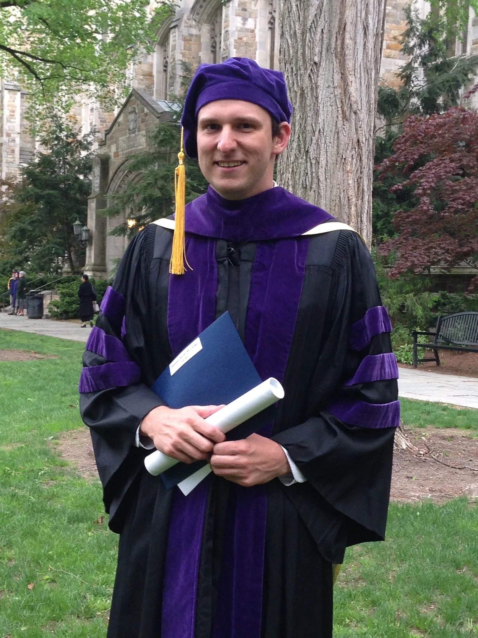 A man in a purple and black graduation gown and cap standing outside near a tree, holding a diploma, a blue folder, and a rolled-up document, with a stone building and other graduates in the background.