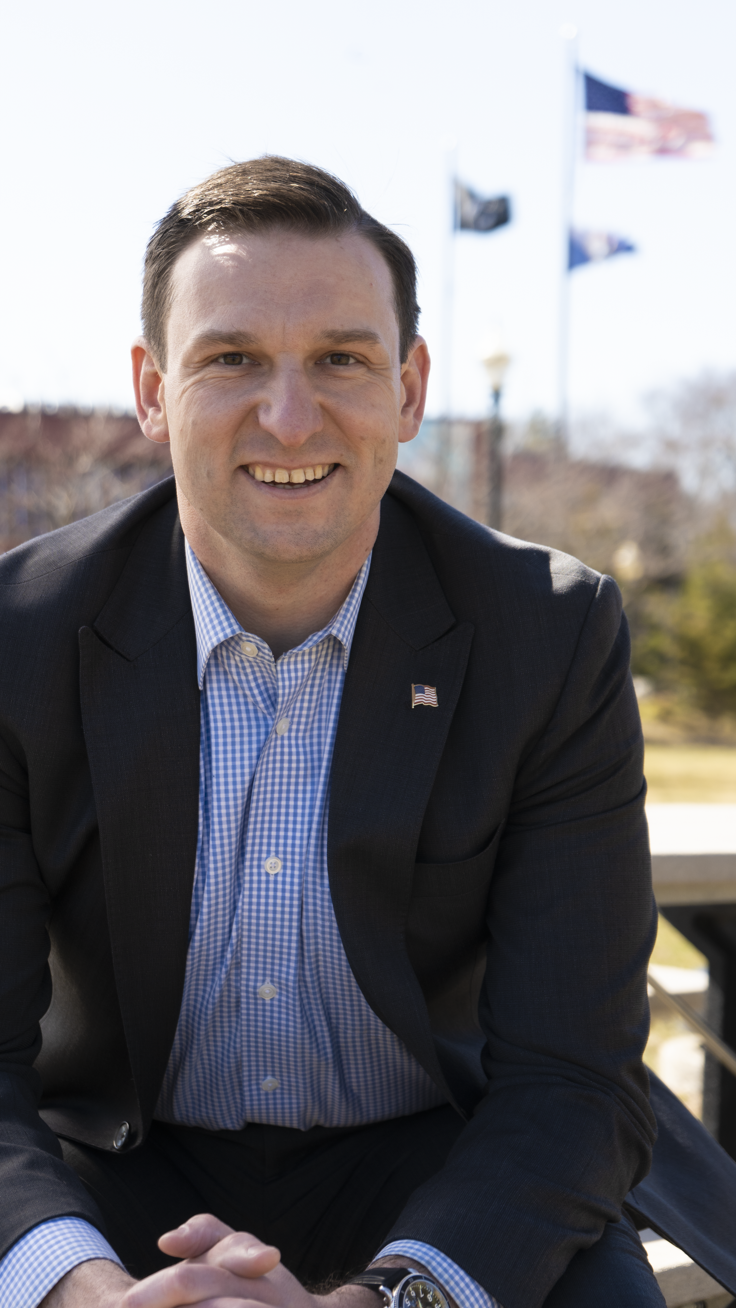 A smiling man in a black blazer and blue checkered shirt, sitting outdoors with American flags in the background.