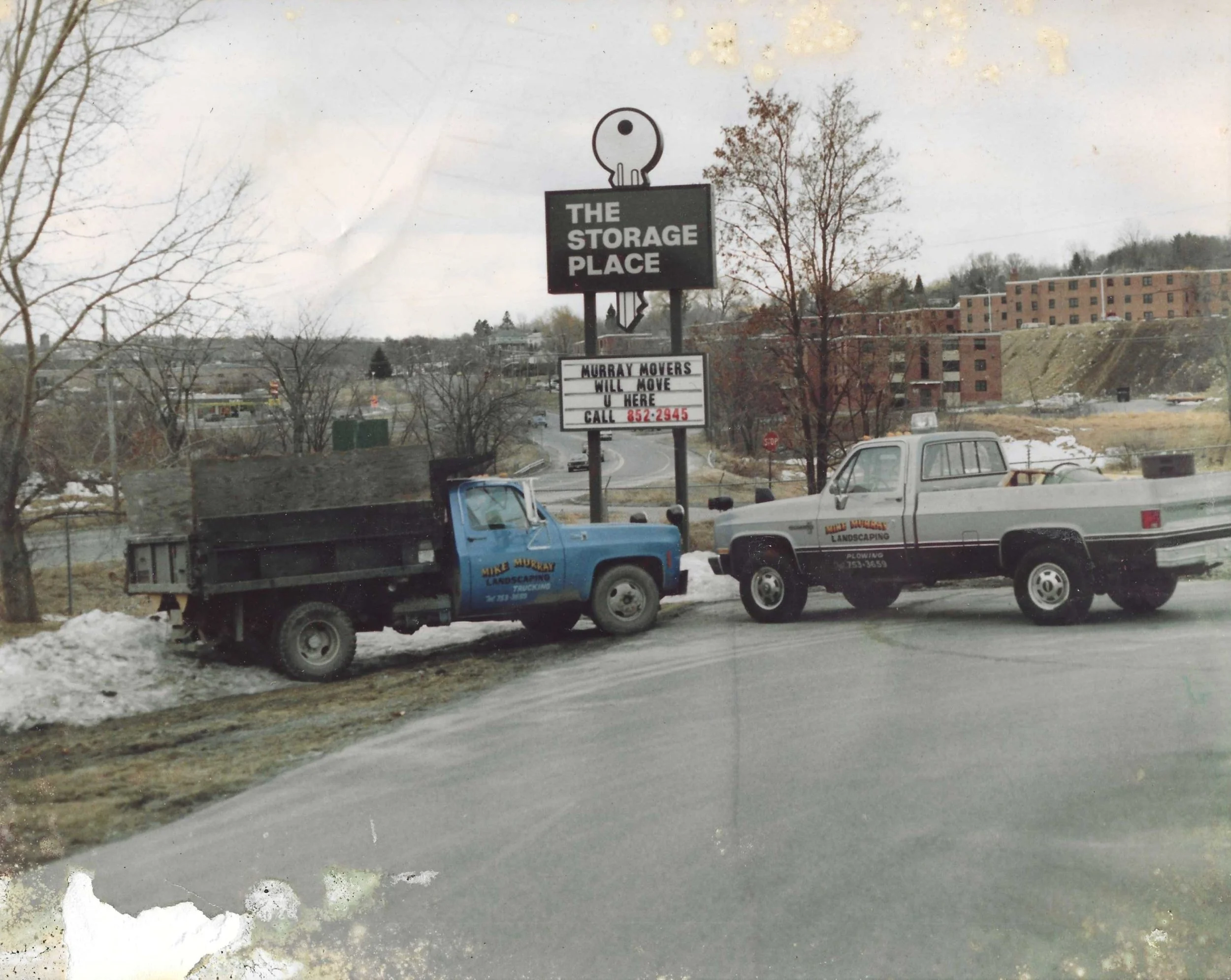 A highway scene with two trucks parked on the side, one blue and one gray, and a sign that reads 'The Storage Place' with a smaller sign below advertising Murray Movers.