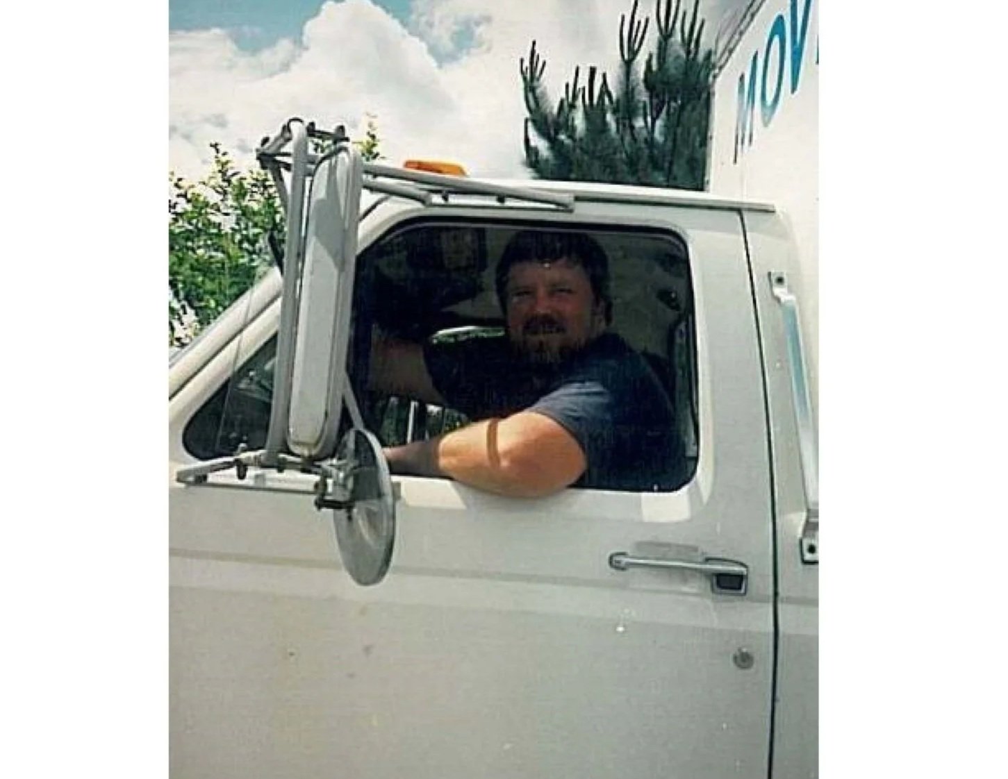A man with a beard and dark hair driving a white truck, looking out the window with his arm resting on the windowsill, under a partly cloudy sky.