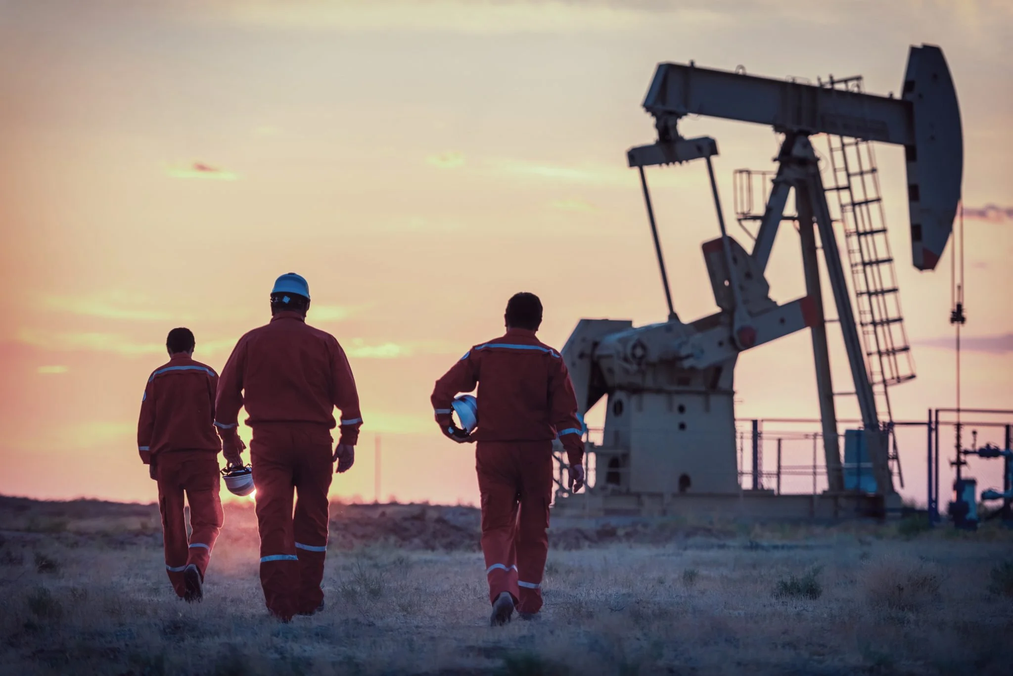 Three workers in red uniforms walking towards oil pumpjack at sunset.