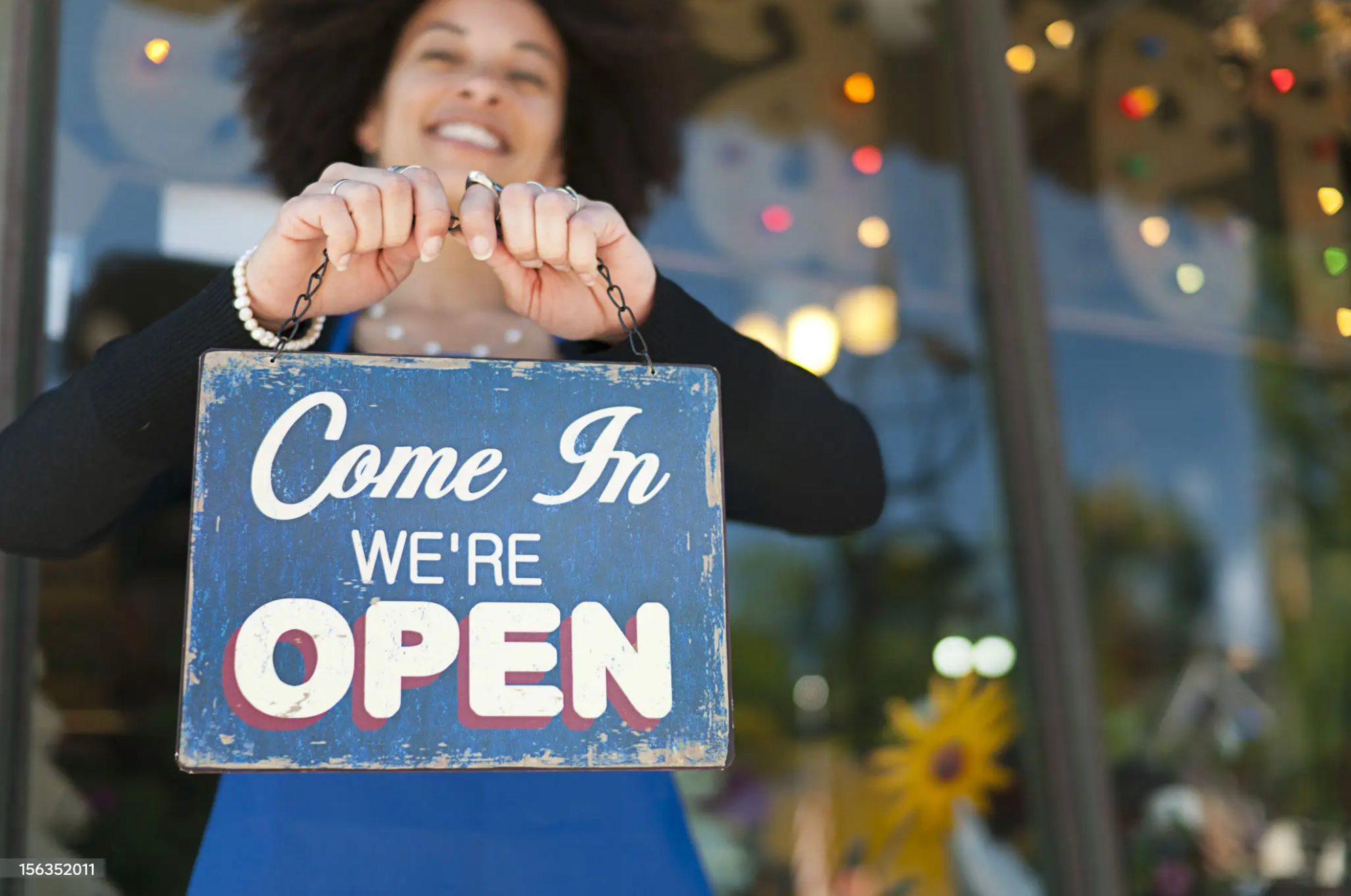 A woman is holding a blue sign that says 'Come In We're OPEN' in front of a storefront decorated with colorful lights.