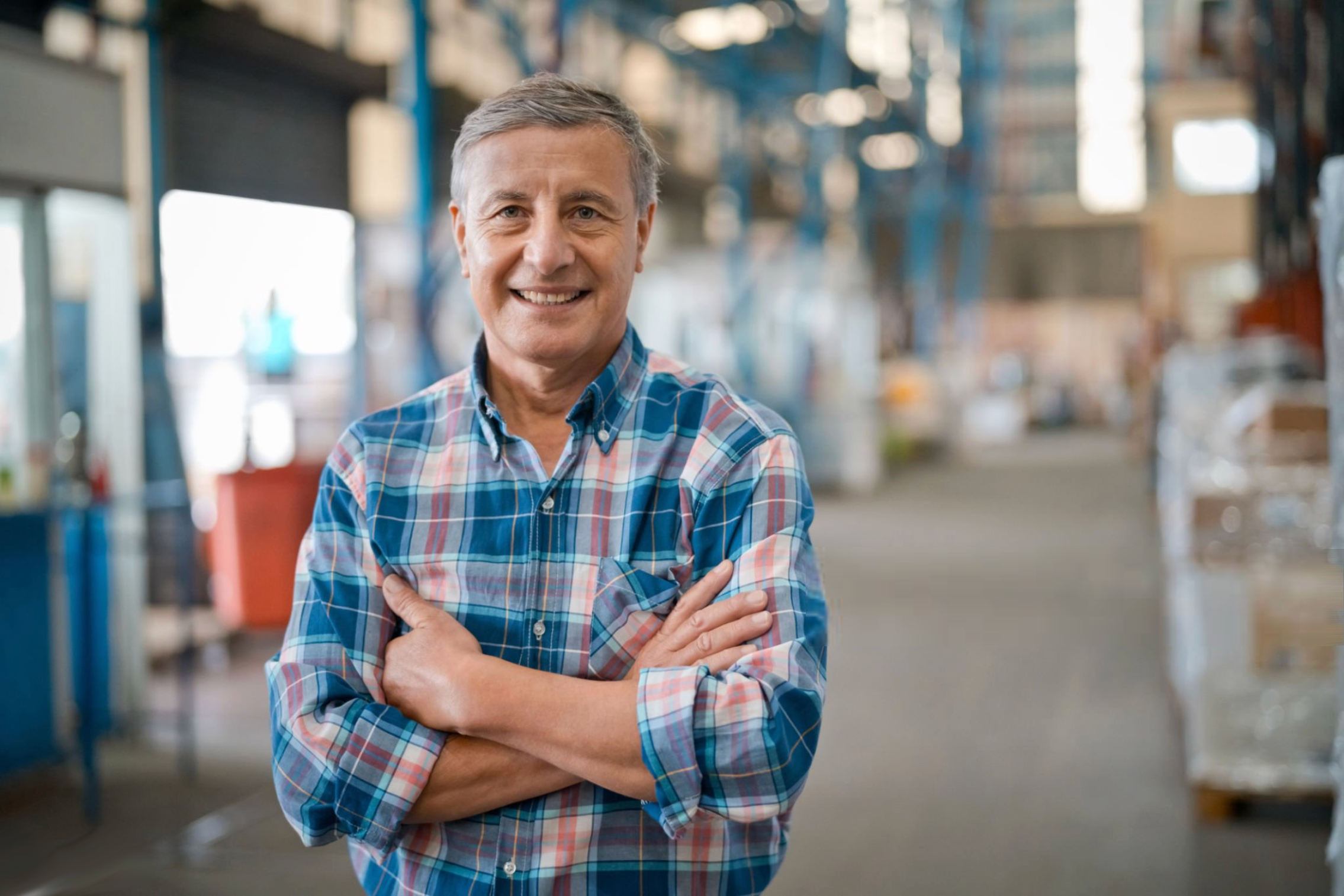 A smiling middle-aged man with gray hair, wearing a blue, red, and white plaid shirt, standing with arms crossed inside a warehouse or industrial space.