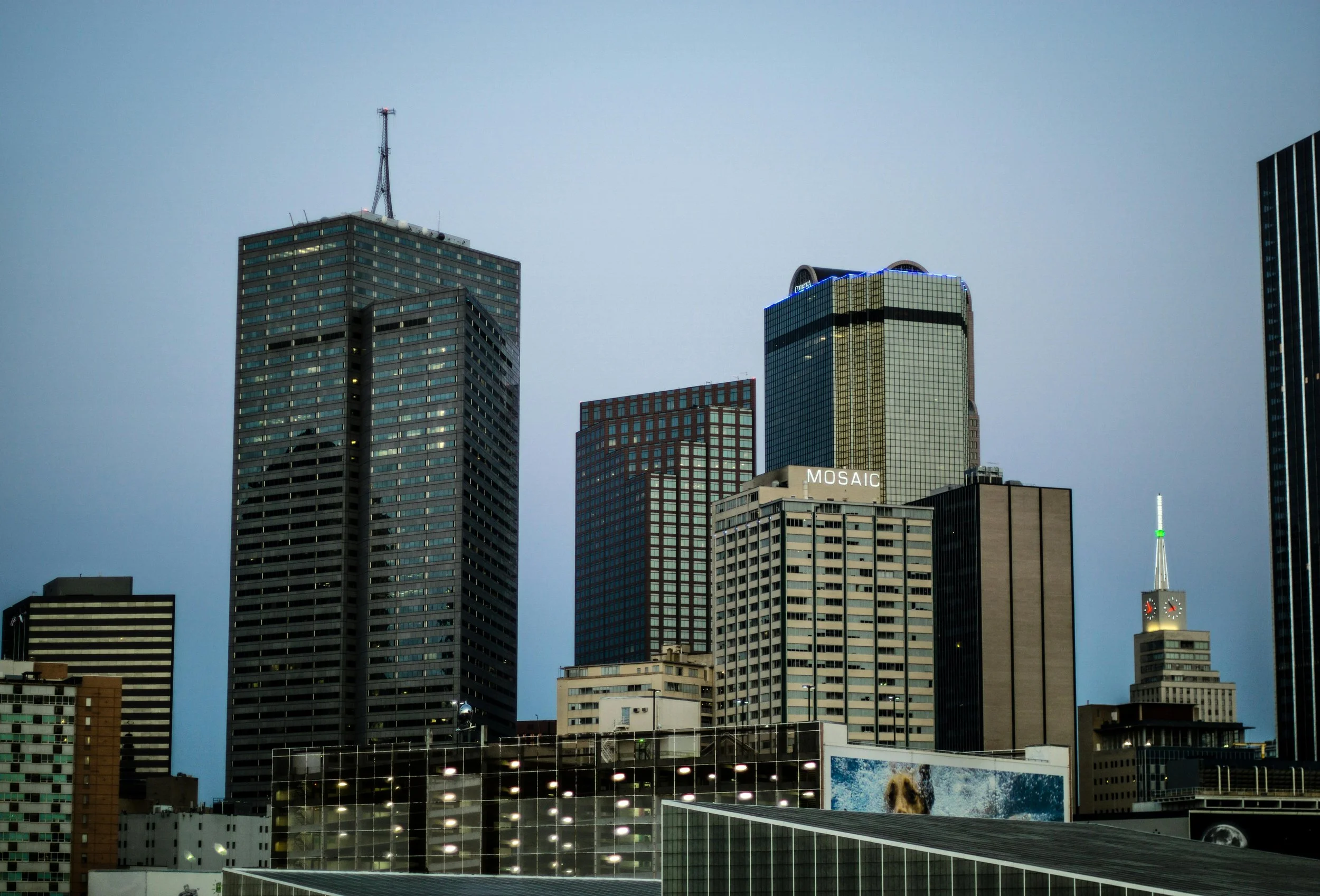 City skyline with tall skyscrapers, including a building labeled 'MOSAIC', and a clock tower with a green-lit spire, under a dusky sky.