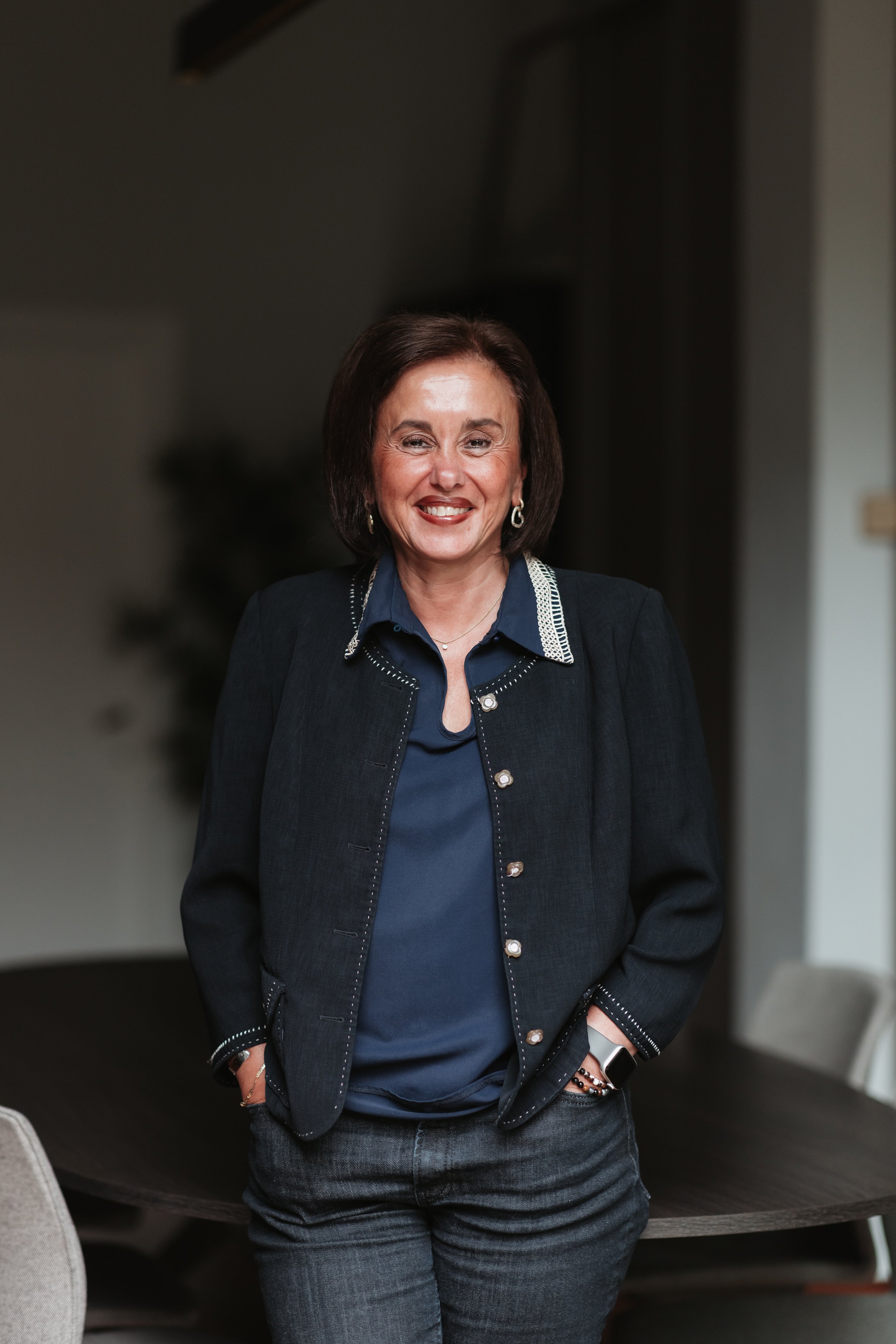 Une femme souriante portant une veste noire et une chemise bleue, avec des accessoires métalliques, debout dans une pièce intérieure.