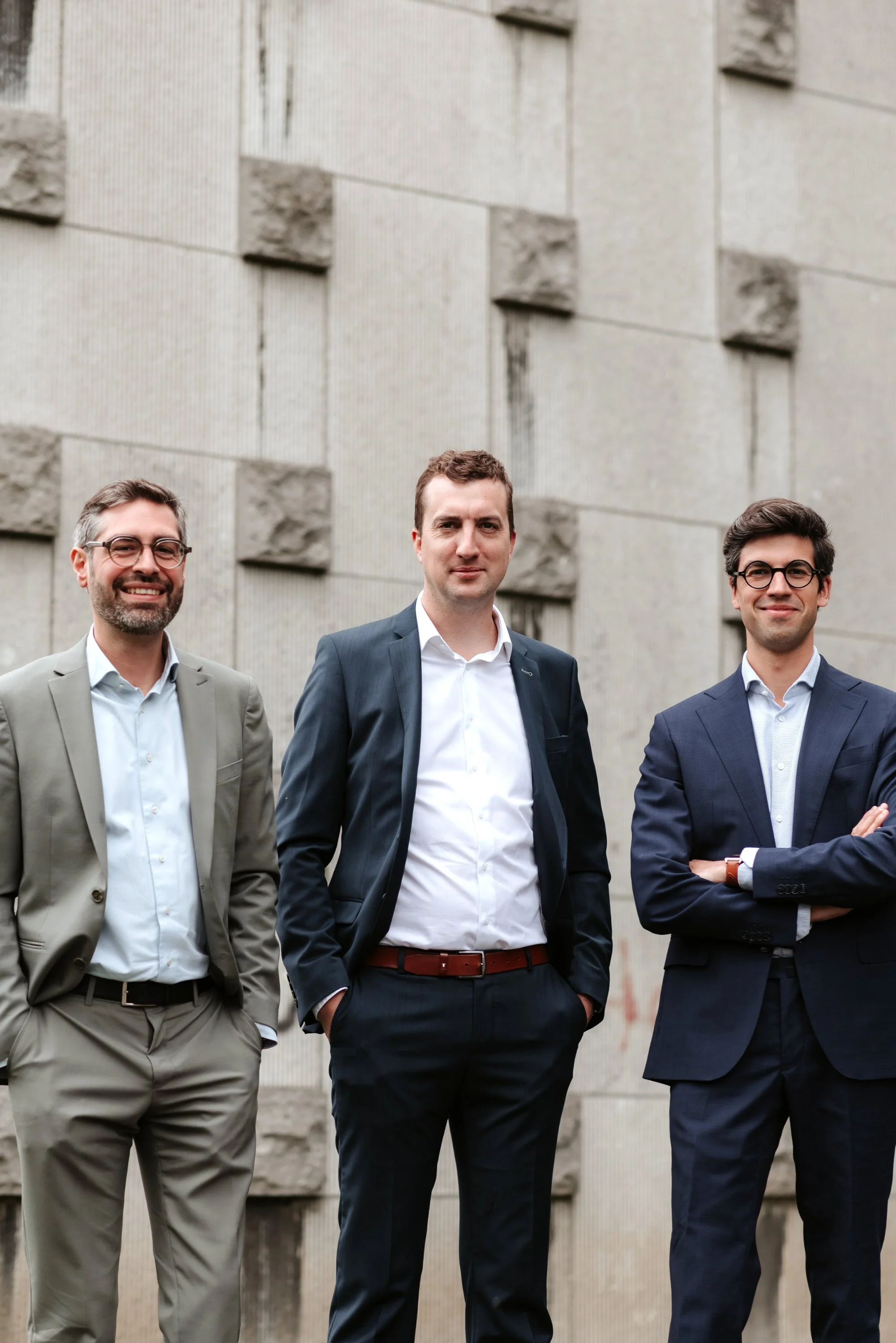 Trois hommes d'affaires en costume posant devant un mur en béton, souriant.