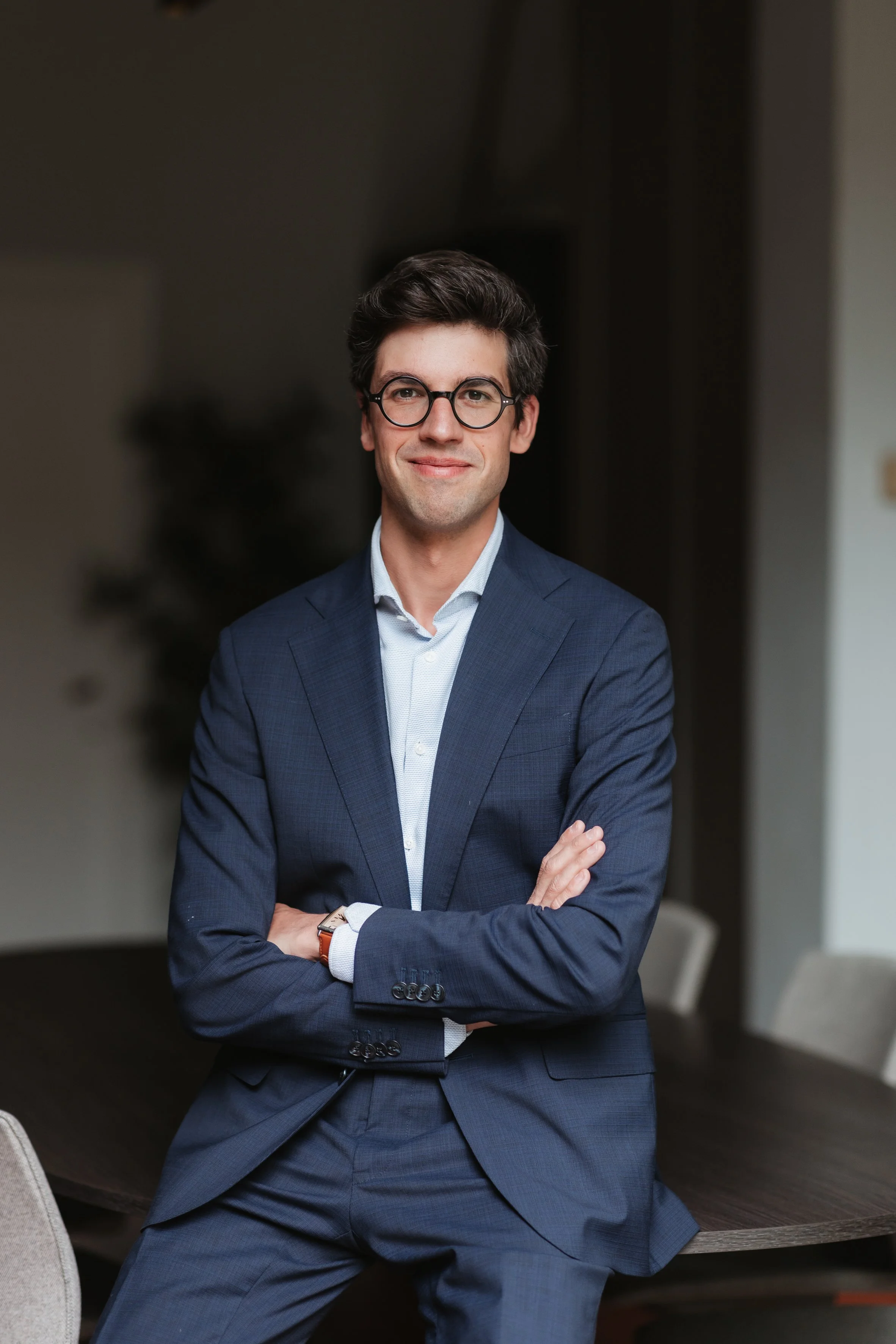 Un homme souriant et portant des lunettes, en costume bleu, avec les bras croisés, dans un intérieur professionnel.