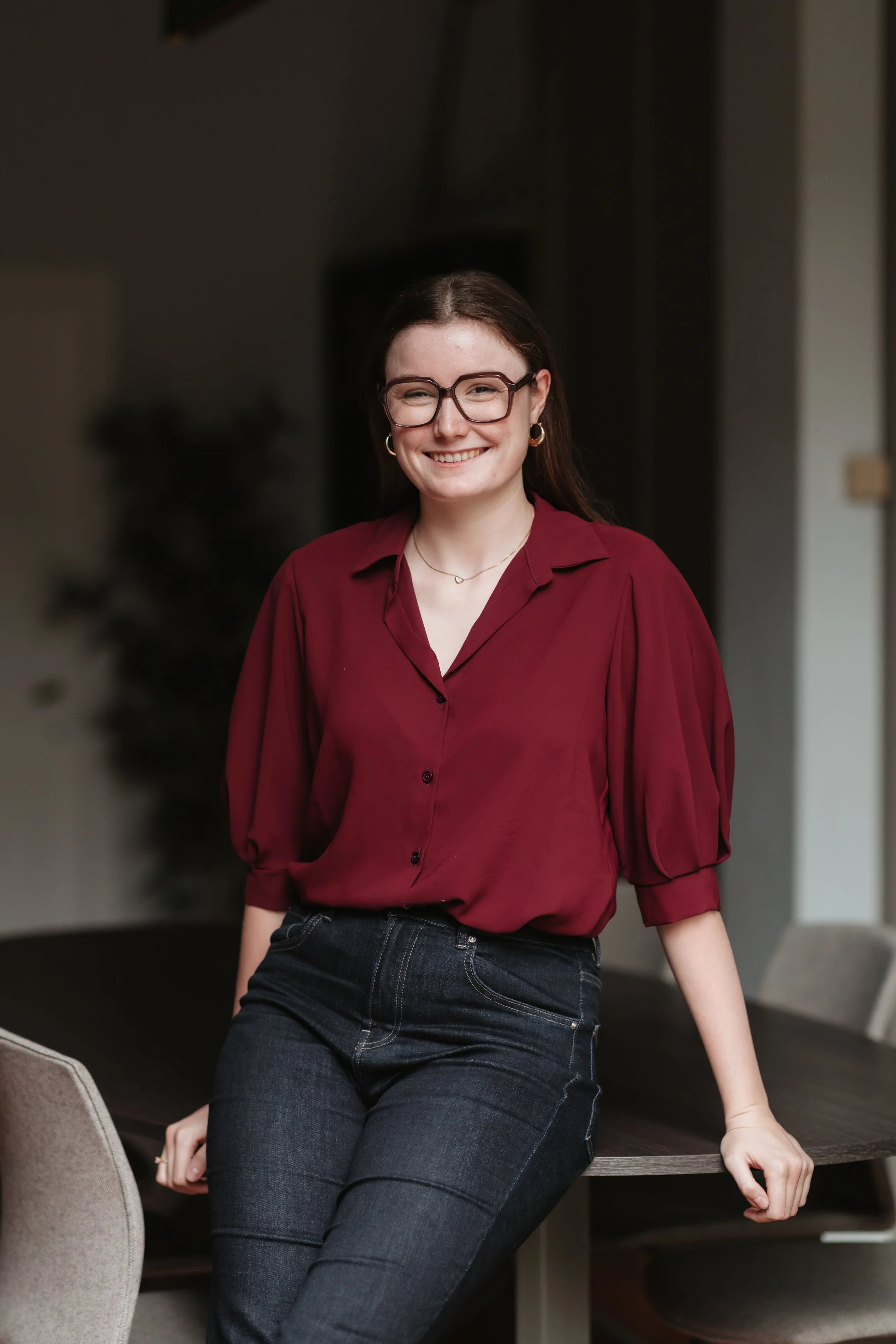 Une femme souriante portant des lunettes, une chemise rouge et un jean, assise sur une table dans un intérieur moderne.
