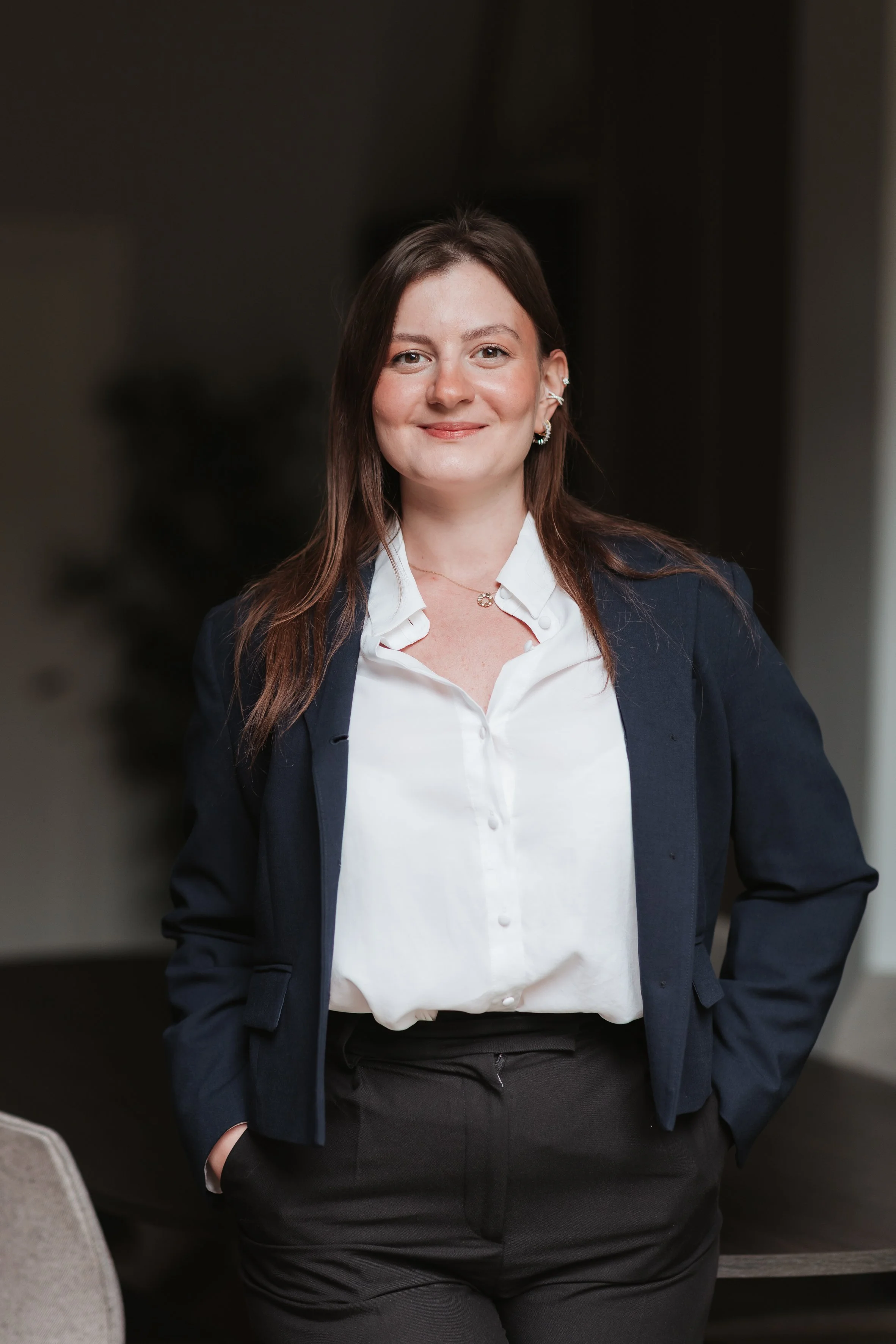 Femme souriante portant un blazer foncé, une chemise blanche et un pantalon noir, debout devant un fond sombre, dans un environnement professionnel.