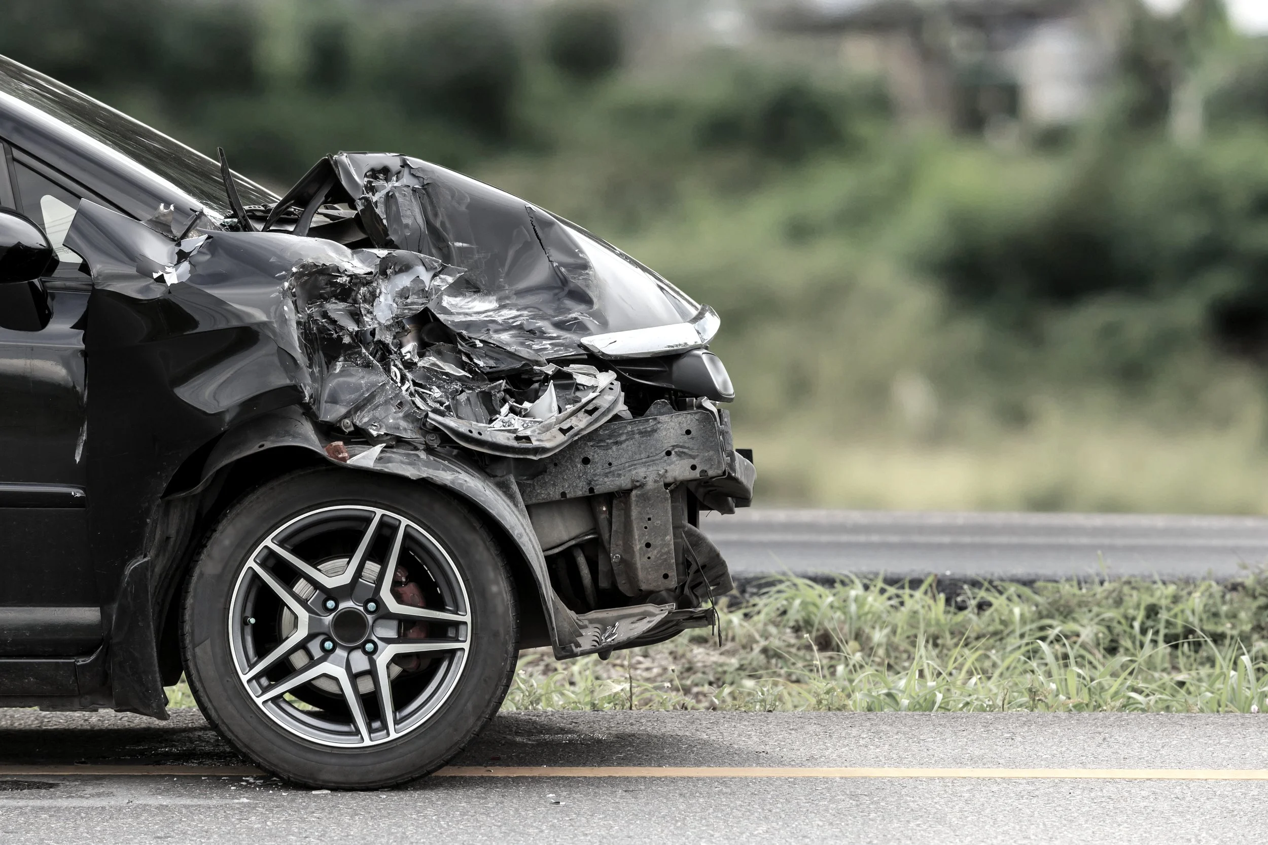Voiture noire endommagée avec une carrosserie écrasée et un pare-chocs avant cassé, stationnée sur le bord de la route, dans un environnement végétal.