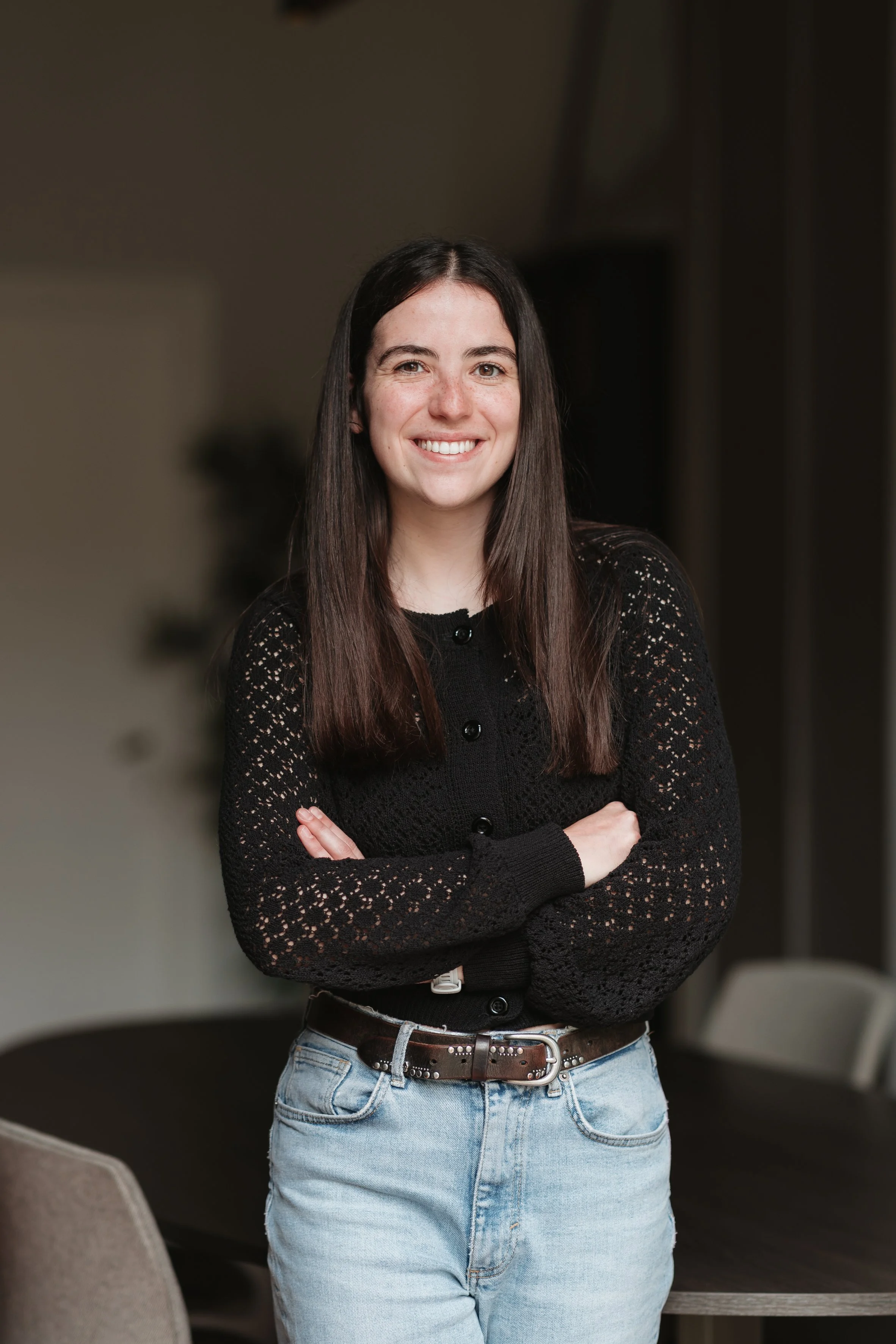 Une jeune femme souriante avec de longs cheveux bruns, portant un pull noir en dentelle et un jean clair, posant avec les bras croisés dans une pièce intérieure.