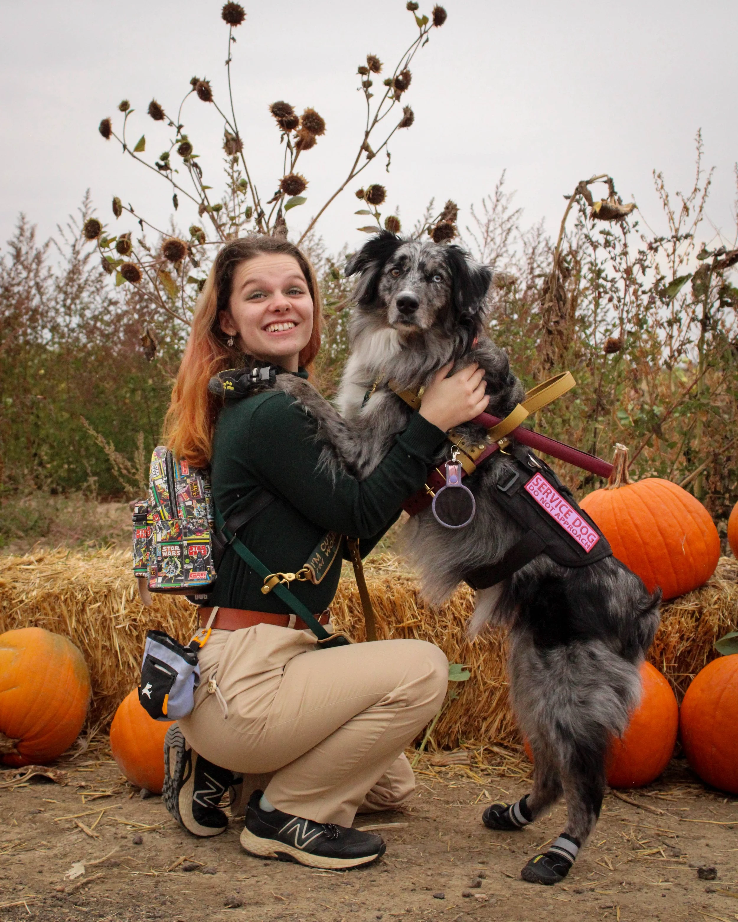 Mara and her dog, Bosun, pose in a pumpkin patch. Mara is kneeling down so Bosun can rest his arms on her shoulders, giving her a hug.