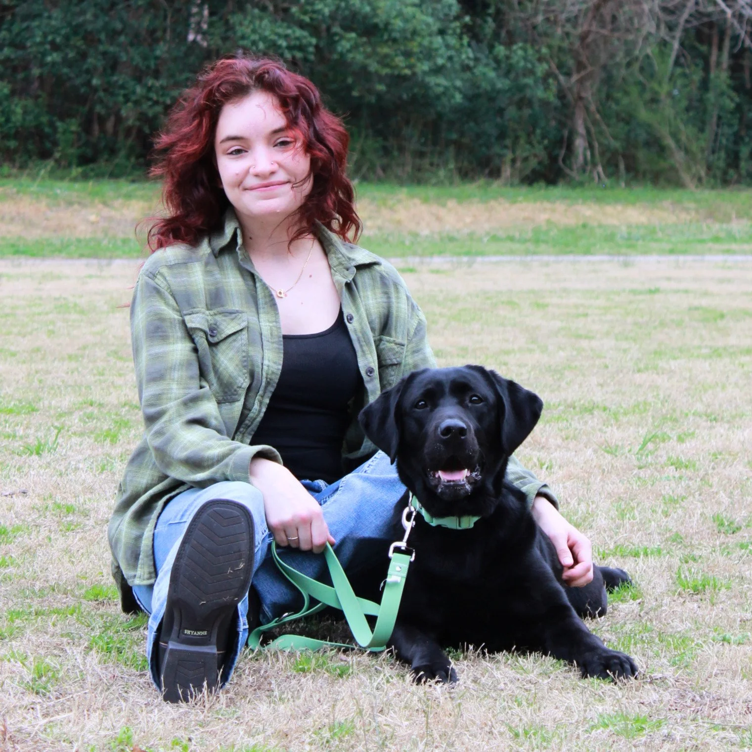 Devin and Ivy sit in a grass field together, smiling, in front of a forest. Devin has wavy red hair, and is wearing a green plaid flannel, black tank top, light colored jeans, and cowboy boots. Ivy is wearing a light green collar and leash.