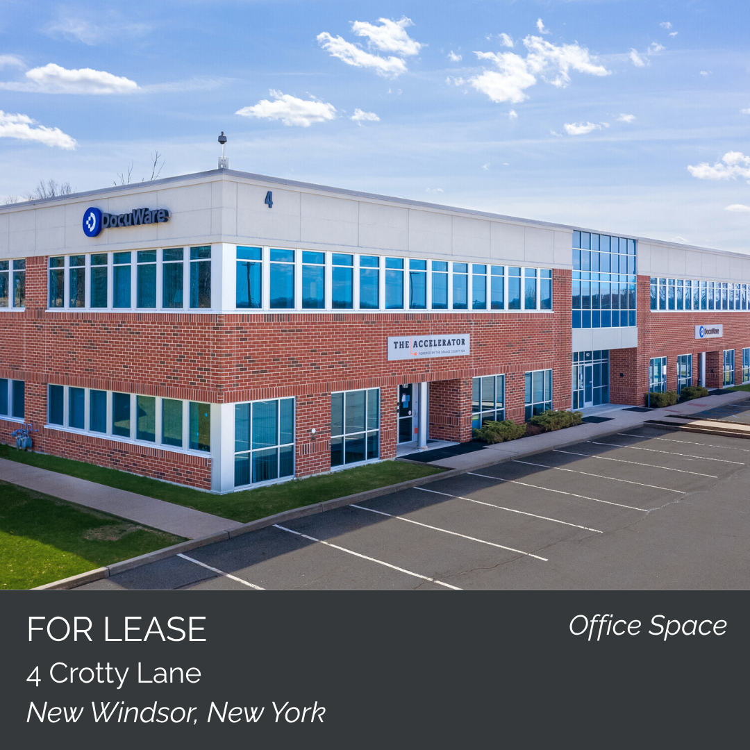 Exterior view of a commercial office building with large blue-tinted windows, red brick walls, and a sign that reads 'The Accelerator.' The building has a parking lot in front and is located in New Windsor, New York.