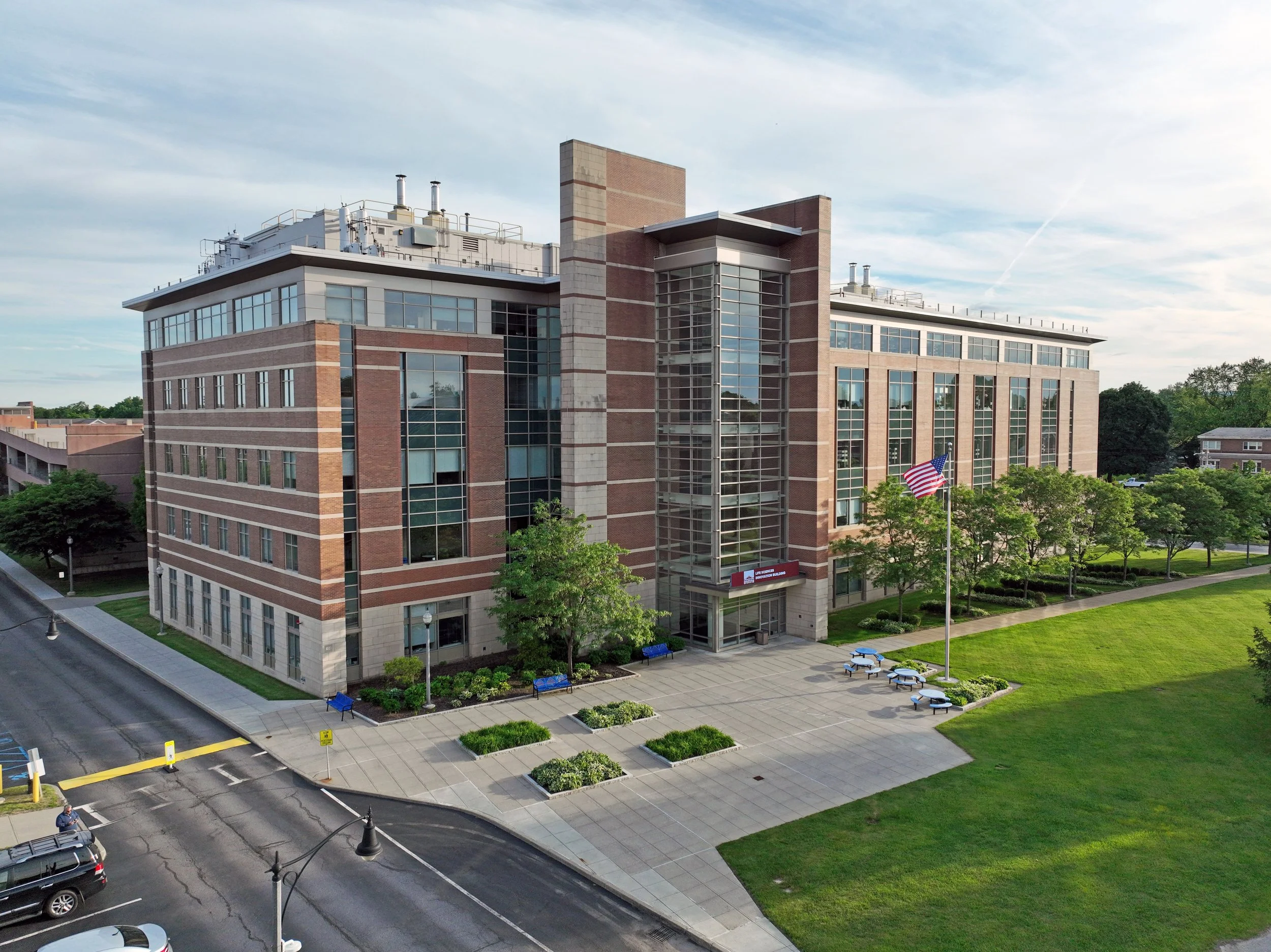 A multi-story brick and glass building with a flagpole flying the American flag, surrounded by trees and a small park area with benches. The sky is partly cloudy.